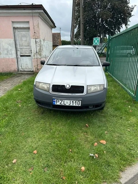 Srebrny samochód marki Skoda zaparkowany na trawniku przed różowym budynkiem z białymi drzwiami. Po prawej stronie znajduje się zielony metalowy płot. Samochód ma polską tablicę rejestracyjną o numerze PZL J110.