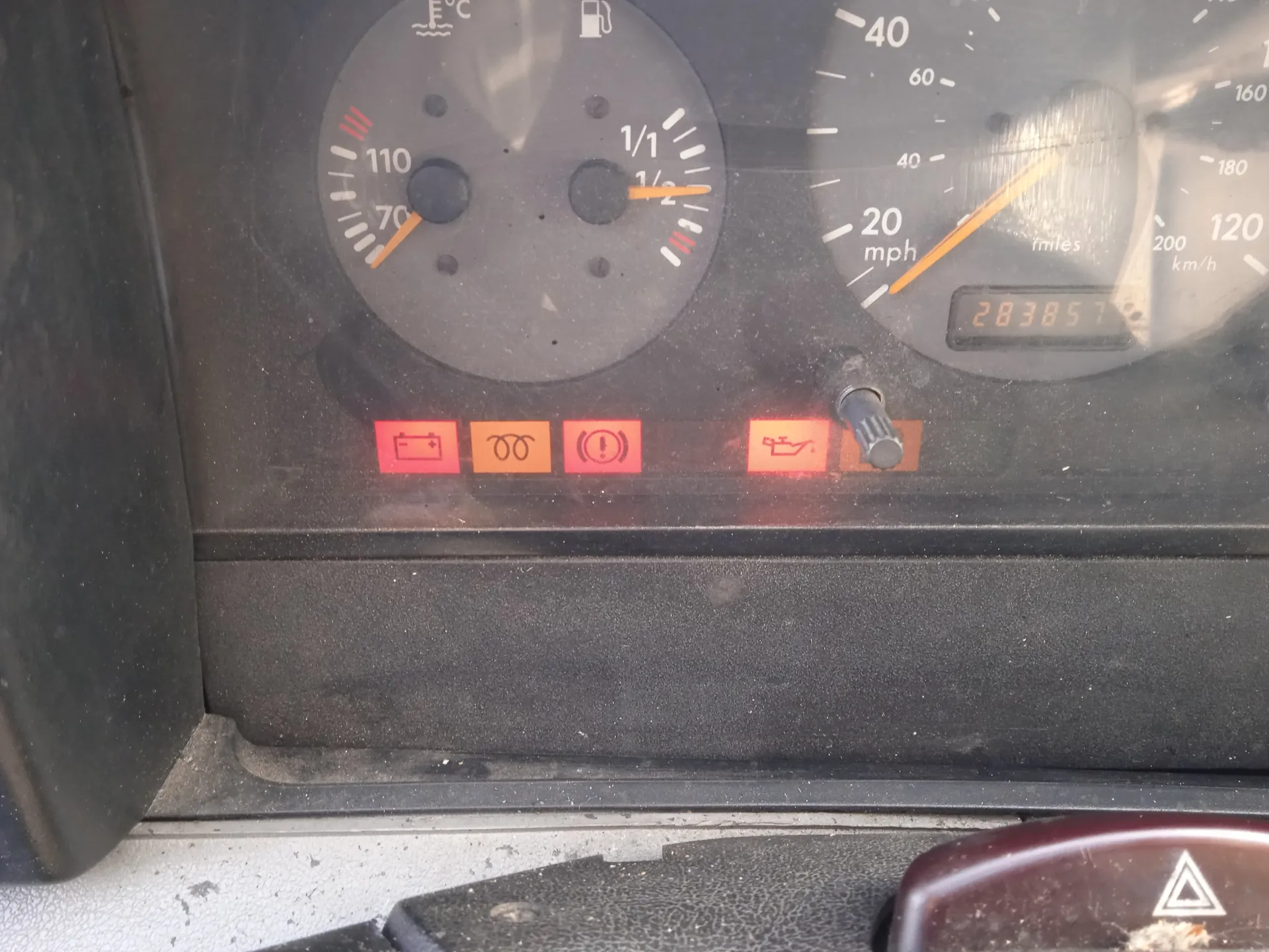 Zbliżenie na deski rozdzielcze samochodu. Po lewej stronie wskaźnik temperatury chłodniczki z zakresem 70-110 i igłą wskazującą około 70, obok wskaźnik paliwa z oznaczeniami 1/1 i 1/2, igła wskazuje blisko 1/2. Po prawej stronie prędkościomierz w mph z igłą na 20 mph oraz cyfrowy licznik przebiegu pokazujący 283857 mil. Poniżej czerwone i pomarańczowe lampki ostrzegawcze: akumulator, światła, hamulce, olej. Widoczna część kierownicy i przycisku awaryjnego z trójkątem. Deska rozdzielcza jest zapylona.
