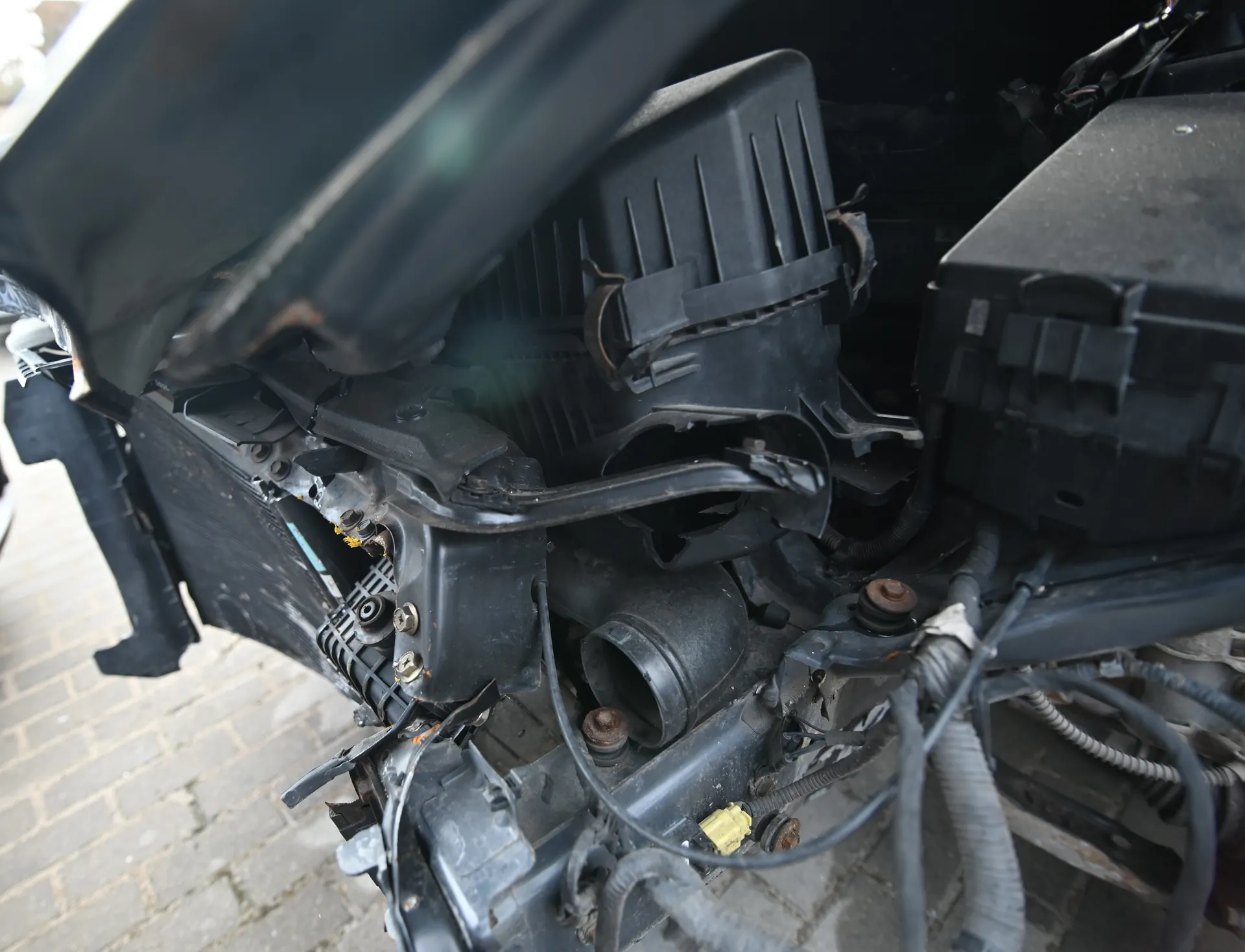 Zbliżenie uszkodzonego przedniego zderzaka i komory silnikowej samochodu. Widoczne są pęknięte czarne elementy plastikowe, przewody elektryczne, metalowe rury oraz żółty złącze. W tle fragment brukowanej nawierzchni. Brak czytelnego tekstu.