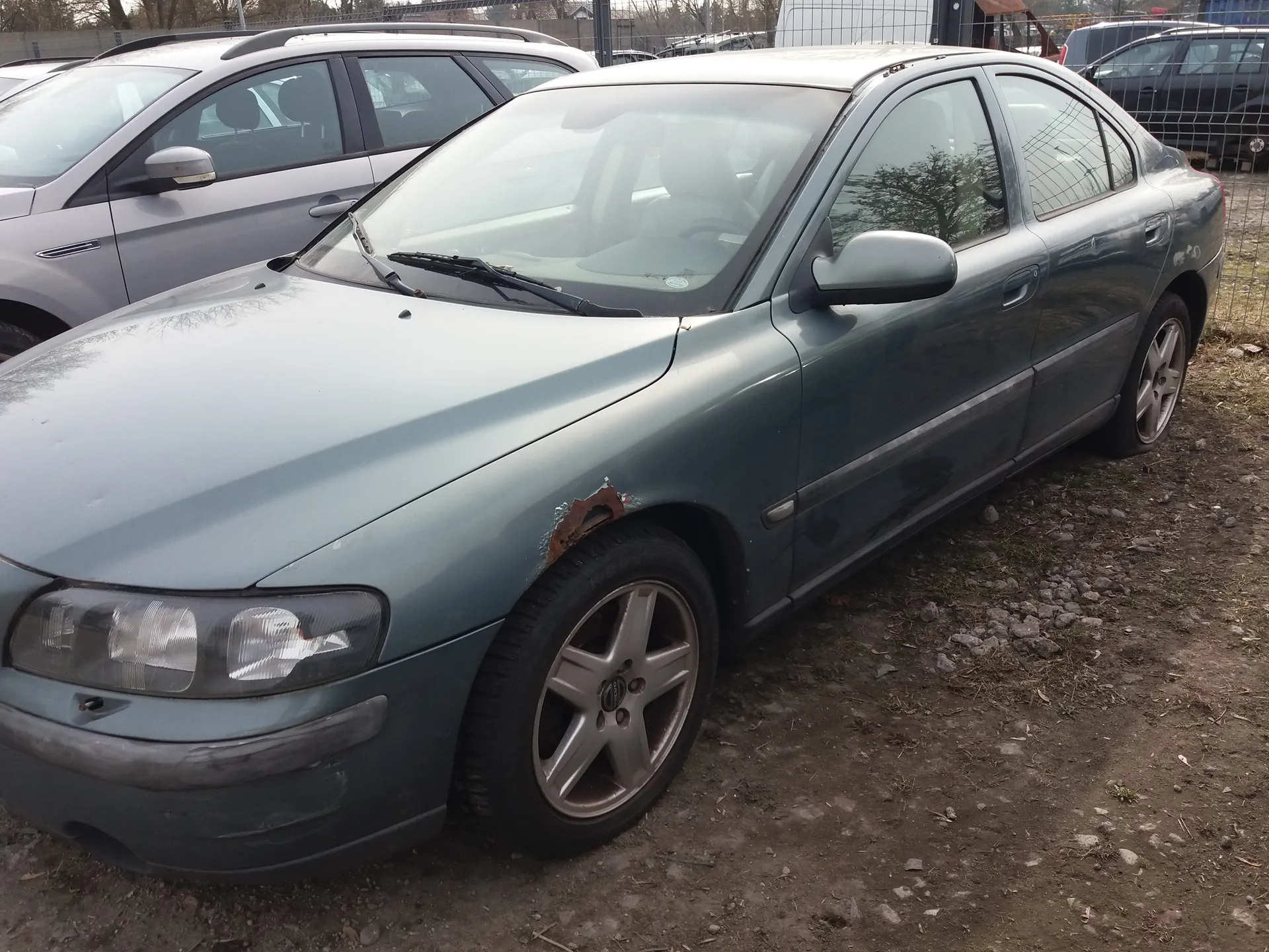 Szary samochód osobowy typu sedan z widocznym uszkodzeniem i rdzą na przednim lewym błotniku. Pojazd stoi na podłożu z żwirem i ziemią. W tle widoczne są inne samochody, metalowe ogrodzenie oraz drzewa. Przednie światła i felgi aluminiowe są czyste, a szyby przeciwsłoneczne na przedniej szybie.