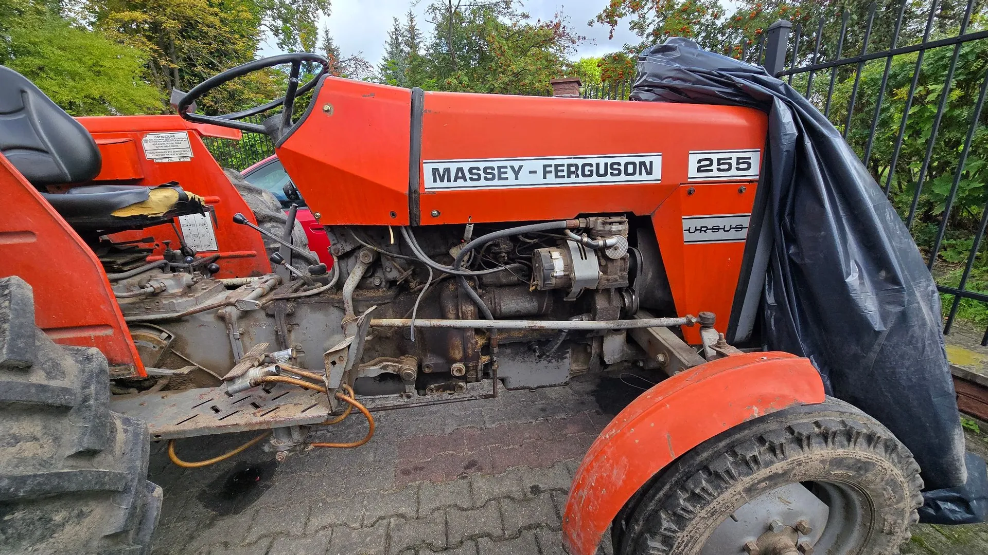 Pomarańczowy traktor marki Massey-Ferguson, model 255. Maszyna jest widoczna z boku, z odsłoniętym silnikiem i układami mechanicznymi. Na masce znajduje się biały napis „MASSEY-FERGUSON 255”, a niżej widnieje napis „URSUS”. Przednia część traktora jest częściowo przykryta czarną folią plastikową. Traktor stoi na kostce brukowej w otoczeniu zieleni.