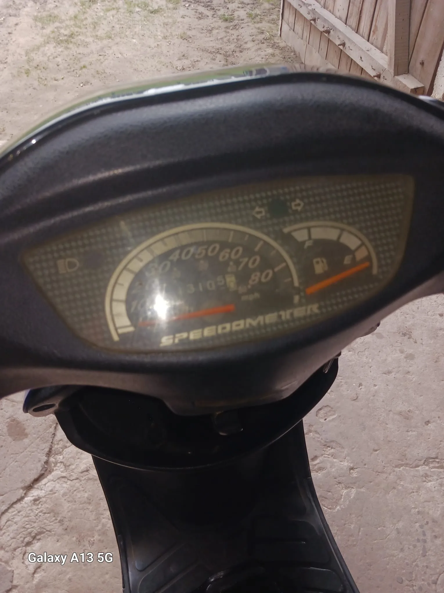 Zbliżenie na czarny wskaźnik prędkości skutera z cyfrowym licznikiem przebiegu 3105. Wskaźnik prędkości pokazuje zakres od 0 do 80 mph, a wskaźnik paliwa znajduje się po prawej stronie. Na dole tablicy widnieje napis SPEEDOMETER. W tle betonowa powierzchnia i drewniane ogrodzenie. W lewym dolnym rogu widoczny napis Galaxy A13 5G.