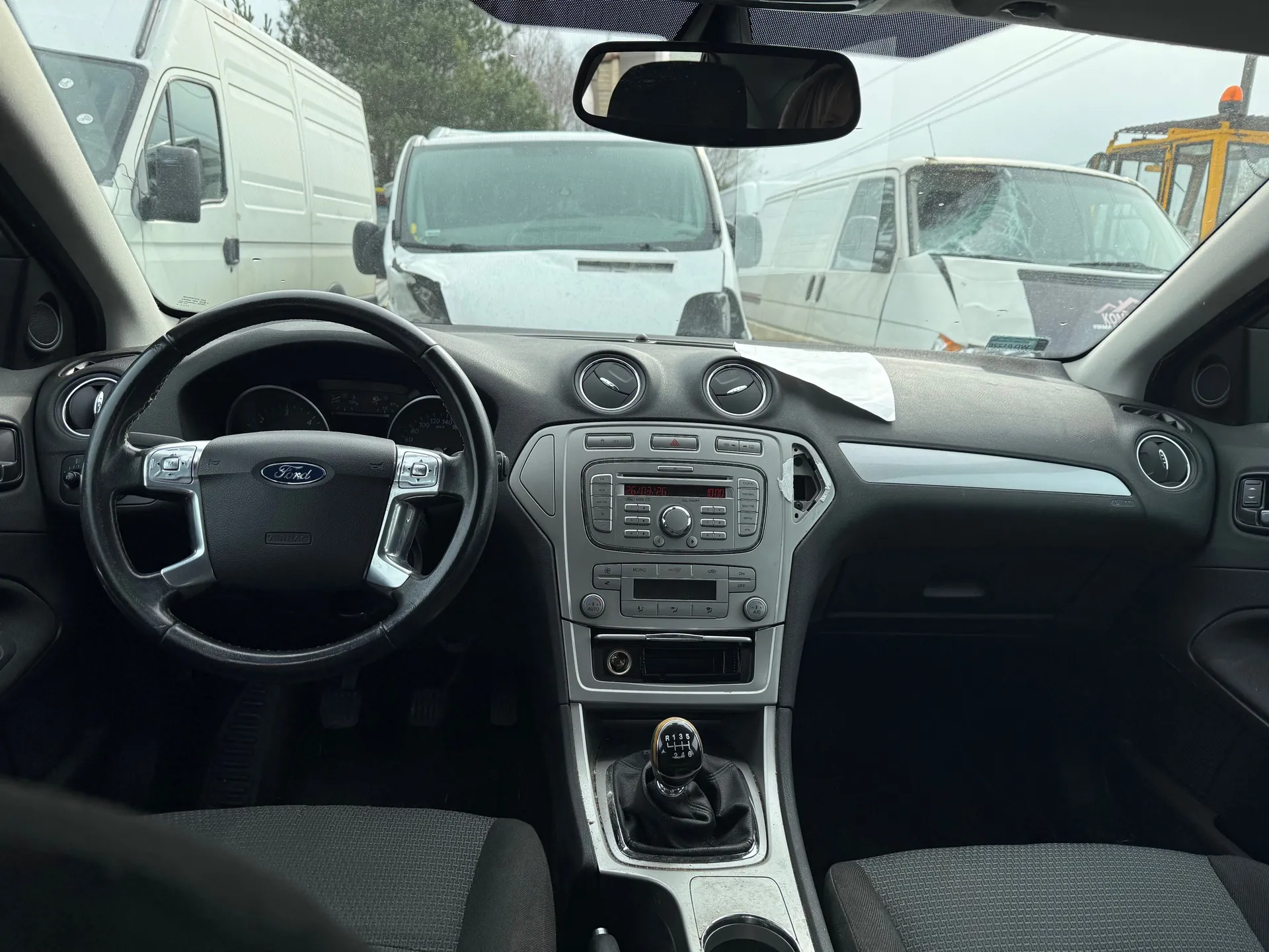 Wnętrze samochodu marki Ford z widocznym kierownicą z logo, deska rozdzielcza z radioodtwarzaczem i przyciskami sterującymi, dźwignią zmiany biegów z oznaczeniami 1-6 i R. Przez przednią szybę widoczne są białe furgony, w tym jeden z pękniętą szybą przednią. Siedzenia pokryte są szarą tkaniną. W lusterku wstecznym widoczny fragment wnętrza pojazdu.