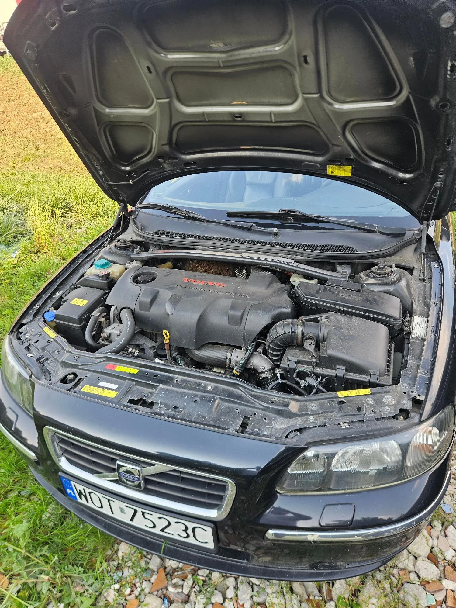 Zdjęcie przodu czarnego samochodu marki Volvo z otwartą maską, ukazującą komorę silnika. Na silniku widnieje napis 'VOLVO'. Samochód posiada polską tablicę rejestracyjną o numerze 'WOT 7523C'. Pojazd stoi na trawiastym podłożu.