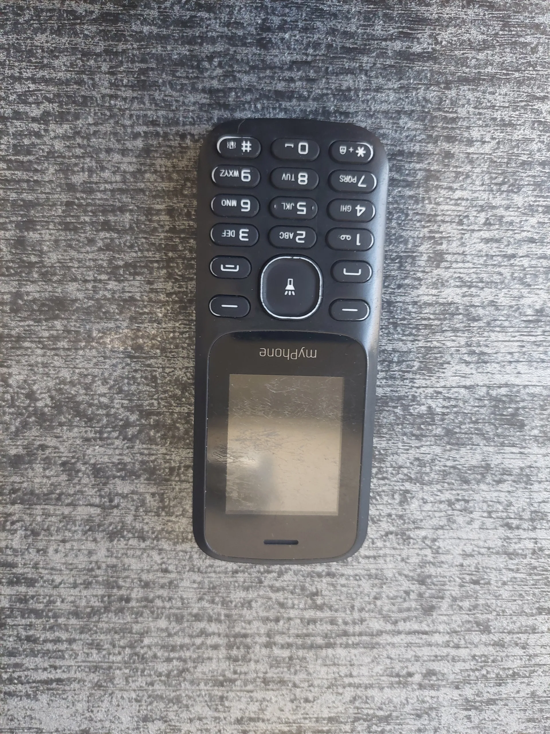 Czarny telefon komórkowy marki myphone z ekranem i klawiaturą. Telefon leży poziomo na szarym, teksturowanym tle. Na klawiaturze widoczne są cyfry od 0 do 9, przyciski funkcyjne oraz symboliki. Na boku ekranu znajduje się napis 'myphone'.