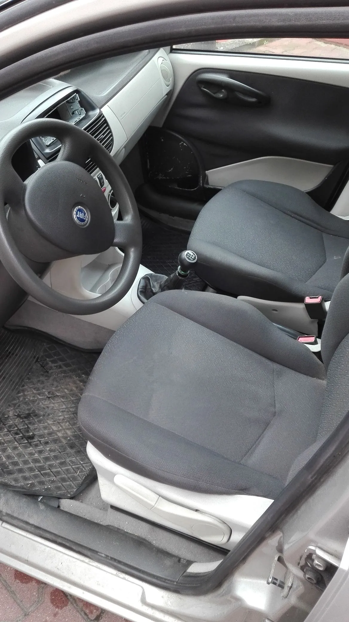 Wnętrze samochodu widziane z perspektywy otwartych drzwi pasażera. Widoczne są dwa przednie fotele z ciemnoszarej tkaniny, kierownica z logo marki Fiat, deska rozdzielcza oraz podłoga z gumowymi dywanikami.