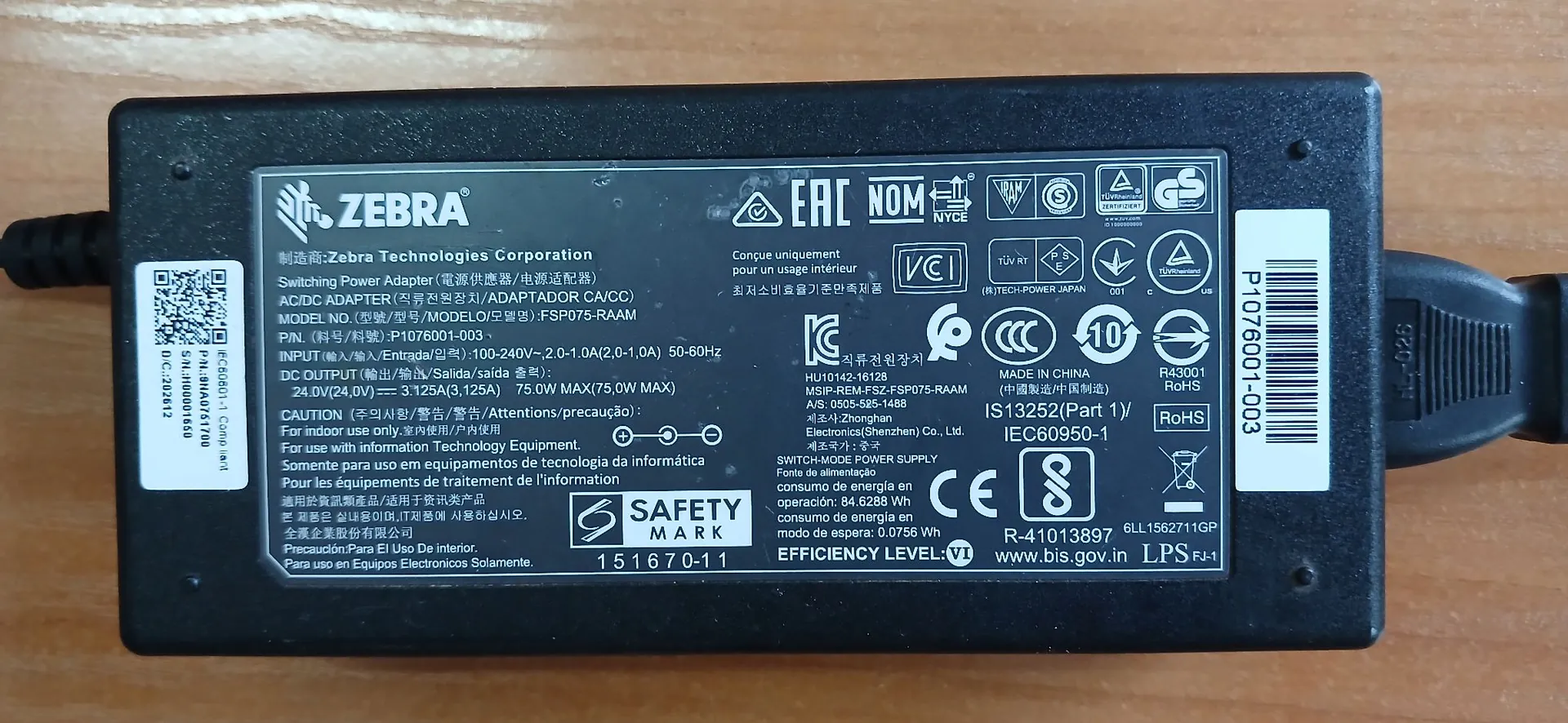 Czarny zasilacz sieciowy marki Zebra. Na obudowie znajduje się biała etykieta z danymi technicznymi, certyfikatami bezpieczeństwa oraz logo producenta. Widoczne napisy obejmują: 'ZEBRA', 'Switching Power Adapter', 'INPUT (输入/entrada/입력): 100-240V~, 2.0-1.0A', 'OUTPUT (输出/salida/출력): 24.0V, 3.125A, 75.0W'. Na etykiecie widnieją liczne symbole certyfikacji, m.in. CE, EAC, UL oraz znak 'SAFETY MARK'.