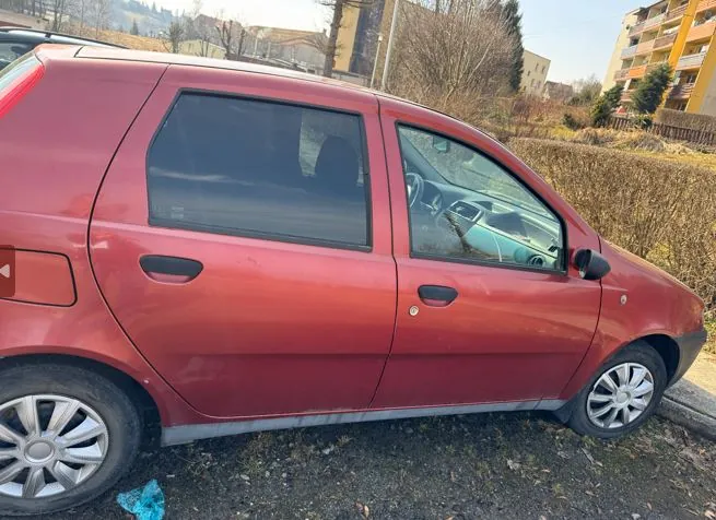 Czerwony samochód osobowy typu hatchback z czterema drzwiami, zaparkowany na podłożu z drobnej kostki. Widoczne są srebrne felgi, ciemne szyby oraz fragmenty otoczenia: krzewy, budynki mieszkalne i drzewa bez liści w tle. Samochód znajduje się na zewnątrz, w słoneczny dzień.