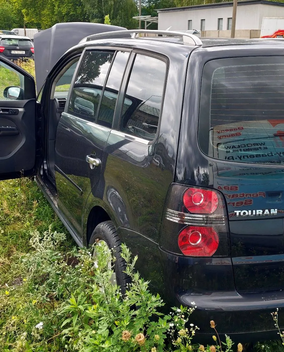 Czarny samochód typu kombi zaparkowany na trawie. Widoczny jest bok i tył pojazdu z otwartymi drzwiami kierowcy oraz otwartą maską silnika. Na tylnej klapie widoczny jest napis TOURAN oraz odbicie w szybie z tekstem, w tym słowem WROCŁAW.