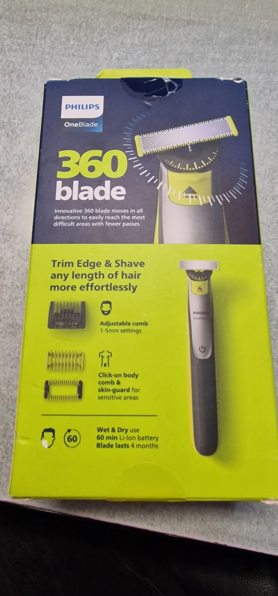 Opakowanie trymera Philips OneBlade 360. Górna część pudełka jest ciemnoniebieska z białym logo PHILIPS i napisem 'OneBlade 360 blade'. Poniżej znajduje się opis: 'Innovative 360 blade moves in all directions to easily reach the most difficult areas with fewer passes'. Dolna część opakowania jest jasnozielona z napisem 'Trim Edge & Shave any length of hair more effortlessly'. Widoczne są ikony i opisy funkcji: 'Adjustable comb 1-5mm settings', 'Click-on body comb & skin-guard for sensitive areas' oraz 'Wet & Dry use, 60 min Li-ion battery, Blade lasts 4 months'. Po prawej stronie znajduje się zdjęcie urządzenia.