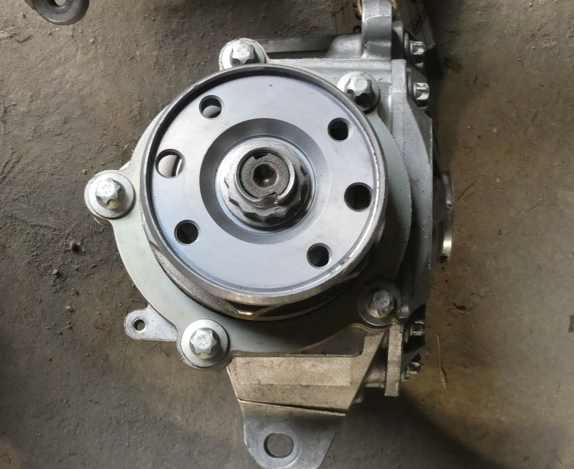 Zbliżenie na metalową część mechaniczną, prawdopodobnie piastę koła lub element układu napędowego, z centralną śrubą i kilkoma otworami montażowymi, zamocowaną do aluminiowej obudowy za pomocą śrub sześciokątnych. Całość spoczywa na szarej, betonowej powierzchni.