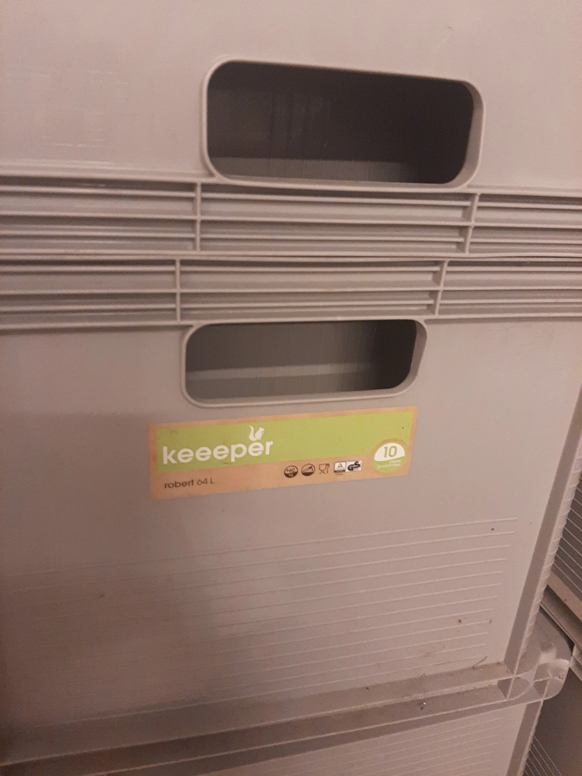 Biały plastikowy pojemnik z pionowymi żebrami i dwoma prostokątnymi otworami po lewej stronie. Na prawej stronie znajduje się etykieta w kolorach zielonym i brązowym z napisem "keeper", cyfrą "10" w okręgu oraz tekstem "robert 64 L". Tekst na etykiecie jest ustawiony pionowo.