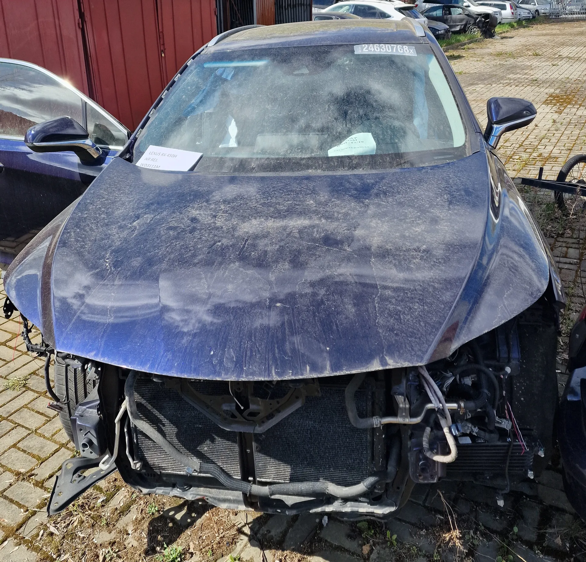 Zniszczony niebieski samochód z usuniętym przodem, odsłaniającym elementy silnika i chłodnicy. Na przedniej szybie widoczny biały znak z numerem 24630768x oraz dwa białe dokumenty na desce rozdzielczej. Samochód stoi na brukowanej powierzchni z trawą między kamieniami. W tle czerwona metalowa brama, inne zaparkowane pojazdy oraz fragment roweru po prawej stronie. Całość pokryta jest kurzem.