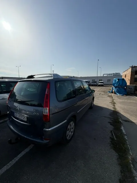 Tylny widok szarego samochodu marki Citroën C8 zaparkowanego na betonowym placu. Samochód ma tablicę rejestracyjną o numerze GU 3H906. W tle widoczne są niskie budynki przemysłowe, inne zaparkowane pojazdy oraz niebieska plandeka przykrywająca jakiś przedmiot. Niebo jest jasne i bezchmurne, z widocznym słońcem w lewym górnym rogu.