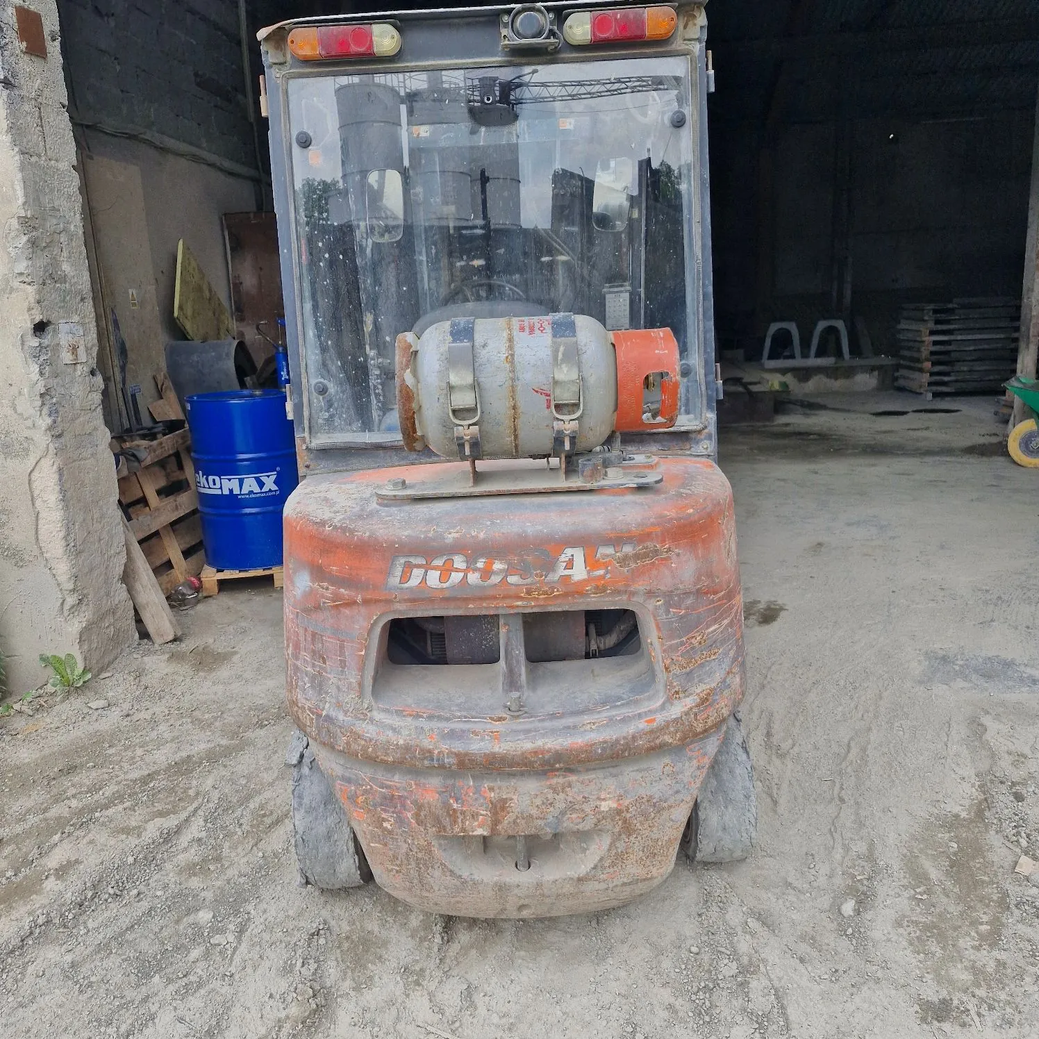 Widok z tyłu pomarańczowego miniladera marki Doosan, zaparkowanego wewnątrz betonowego garażu lub hali. Maszyna jest zakurzona i posiada widoczne ślady użytkowania. Z tyłu pojazdu zamontowany jest szary zbiornik z pomarańczową zakrętką. W tle po lewej stronie widoczna jest niebieska beczka z napisem 'KOMAX' oraz drewniane palety.