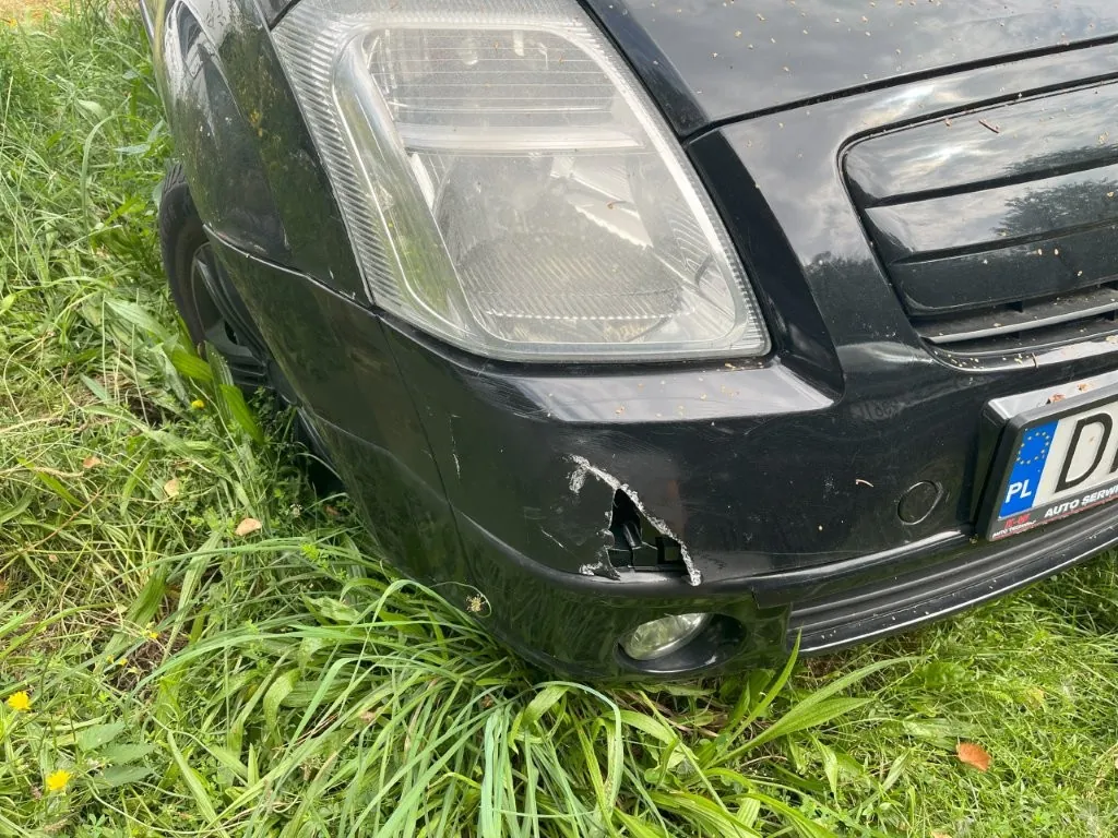 Zbliżenie na przednią część czarnego samochodu zaparkowanego na trawie. Widoczny jest prawy reflektor oraz uszkodzony zderzak z pęknięciem i odpryskiem lakieru. W prawym dolnym rogu widoczny jest fragment tablicy rejestracyjnej z oznaczeniem PL oraz napisem AUTO SERWIS.