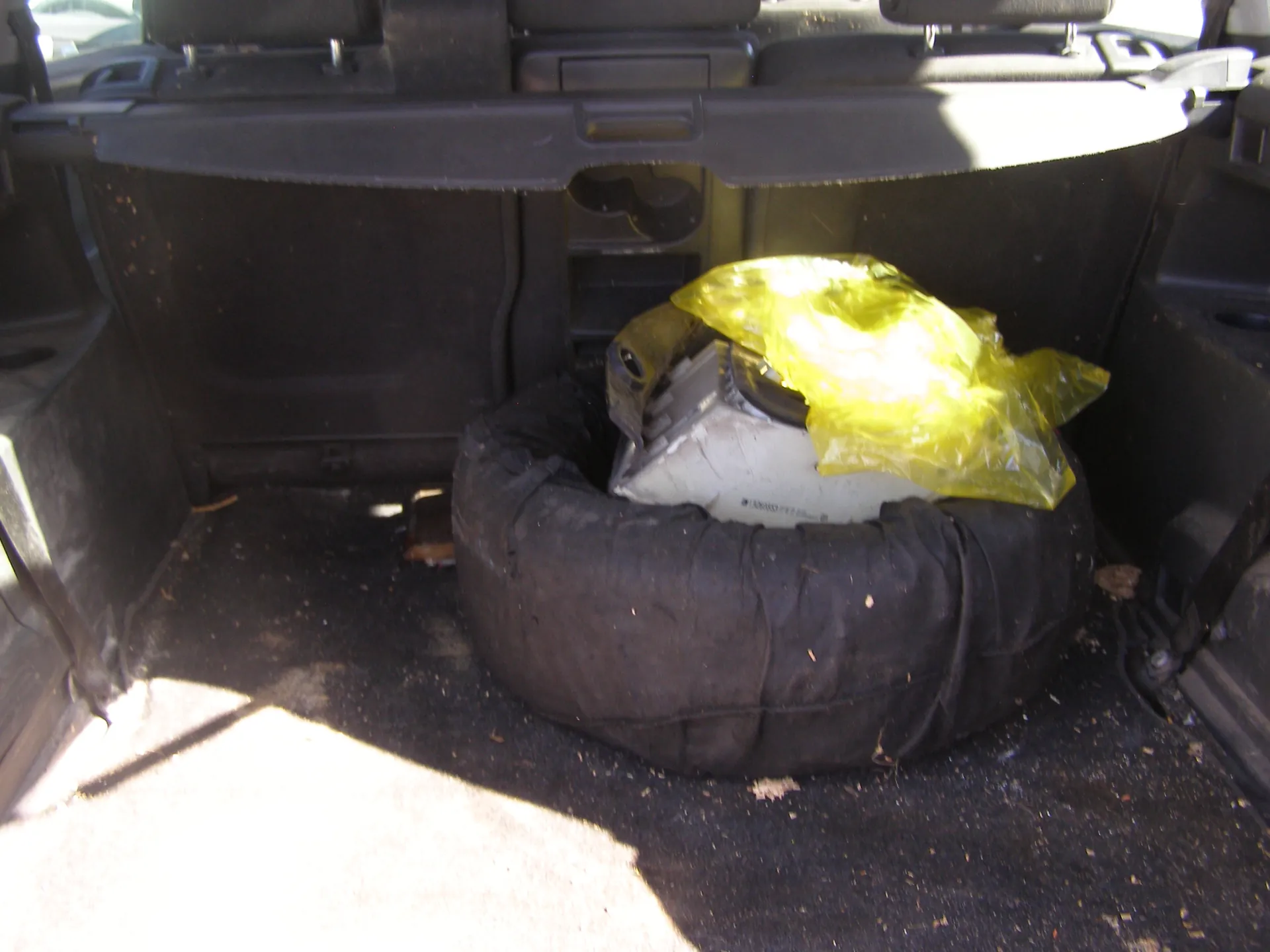 Wnętrze bagażnika samochodu z czarnym kołem zapasowym owiniętym czarną tkaniną. Na kole znajduje się żółta plastikowa torba z białym przedmiotem, na którym widoczny jest nieczytelny tekst. Podłoga bagażnika jest czarna, z widocznymi drobnymi zabrudzeniami i śladami brudu. W tle widoczne są czarne oparcia tylnych siedzeń oraz fragmenty wnętrza pojazdu. Oświetlenie wskazuje na dzień, z widocznymi promieniami słońca wpadającymi do bagażnika.