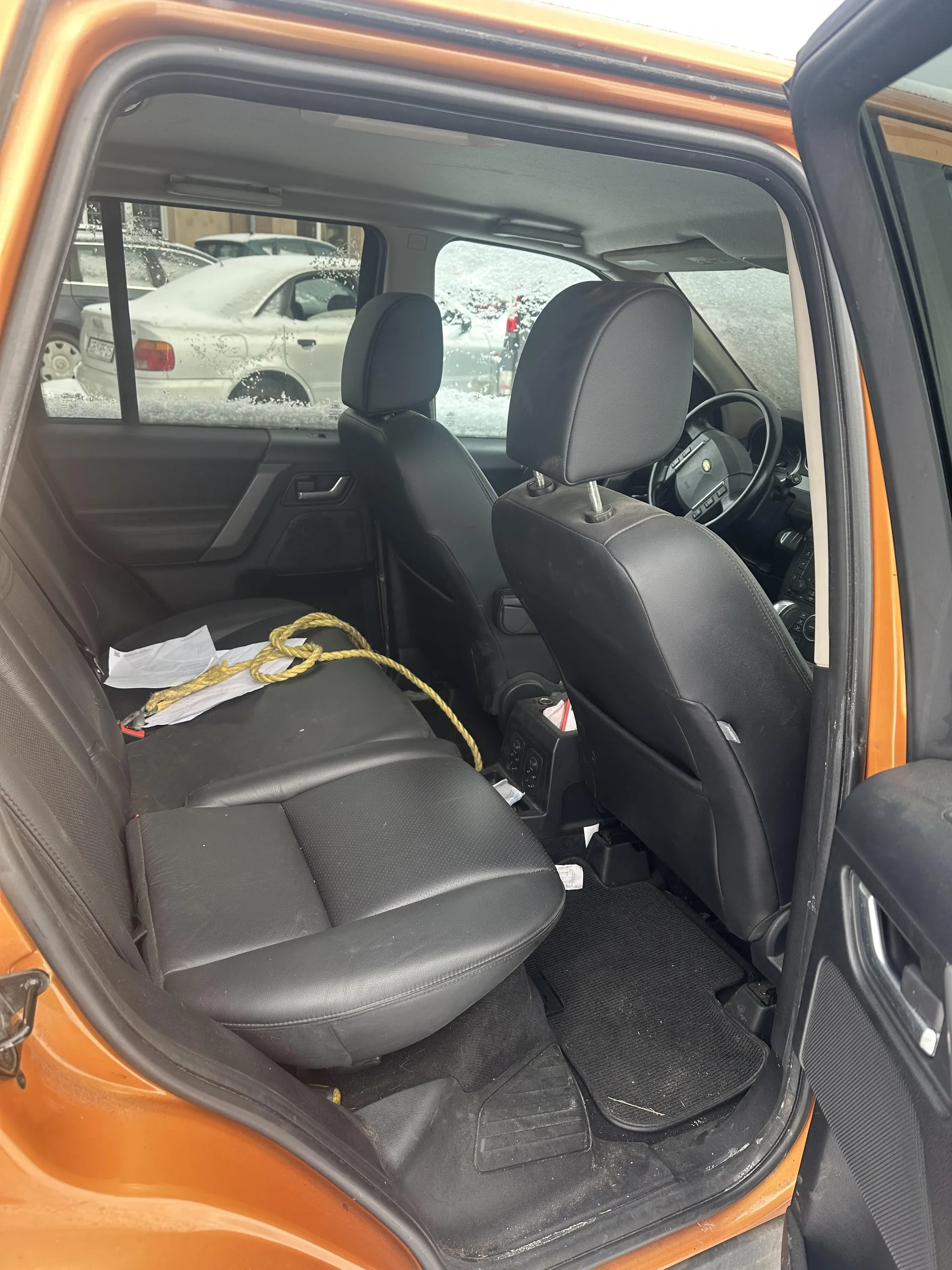 Wnętrze pomarańczowego samochodu z otwartymi drzwiami. Widoczne czarne skórzane siedzenia, na tylnym siedzeniu leży żółta lina i białe dokumenty. Na podłodze czarne dywaniki. Przez okna widać zaparkowane samochody pokryte śniegiem.