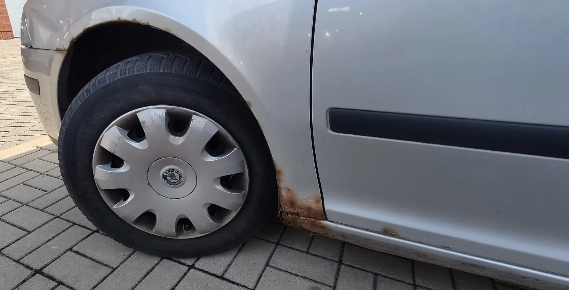 Zbliżenie na przednie lewe koło srebrnego samochodu marki Skoda. Na nadkolu oraz dolnej krawędzi drzwi widoczne są wyraźne ślady rdzy i korozji lakieru. Samochód stoi na szarym kostkowym podjeździe.