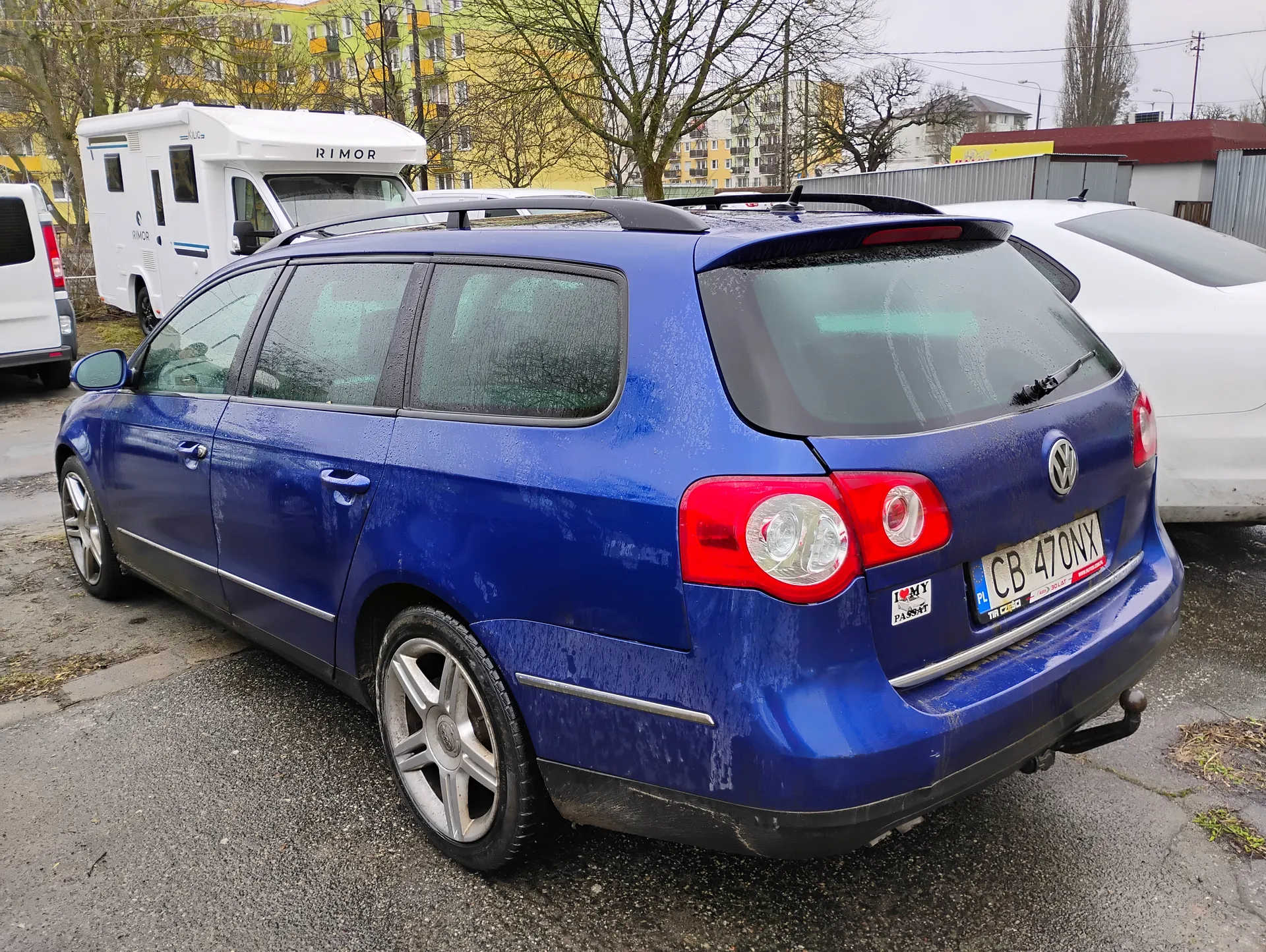 Niebieski samochód osobowy typu kombi marki Volkswagen z tablicą rejestracyjną CB 470NX, zaparkowany na mokrej nawierzchni asfaltowej. Na tylnej szybie widoczny wycieraczka, a na bagażniku naklejka z napisem "I ❤️ MY PASSAT". W tle biały naczepowy pojazd z napisem "RIMOR", żółty blok mieszkalny, drzewa bez liści oraz inne zaparkowane pojazdy. Samochód pokryty jest kroplami wody, a nawierzchnia ulicy jest wilgotna.
