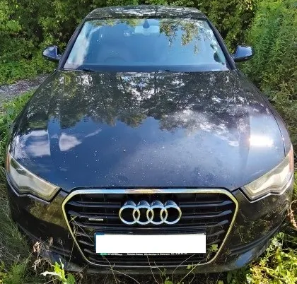 Widok z przodu na czarny samochód marki Audi zaparkowany na trawie przed zielonymi krzewami. Na masce samochodu widoczne są odbicia nieba i drzew. Samochód posiada charakterystyczny grill z czterema splecionymi pierścieniami logo Audi oraz białą, zakrytą tablicę rejestracyjną.