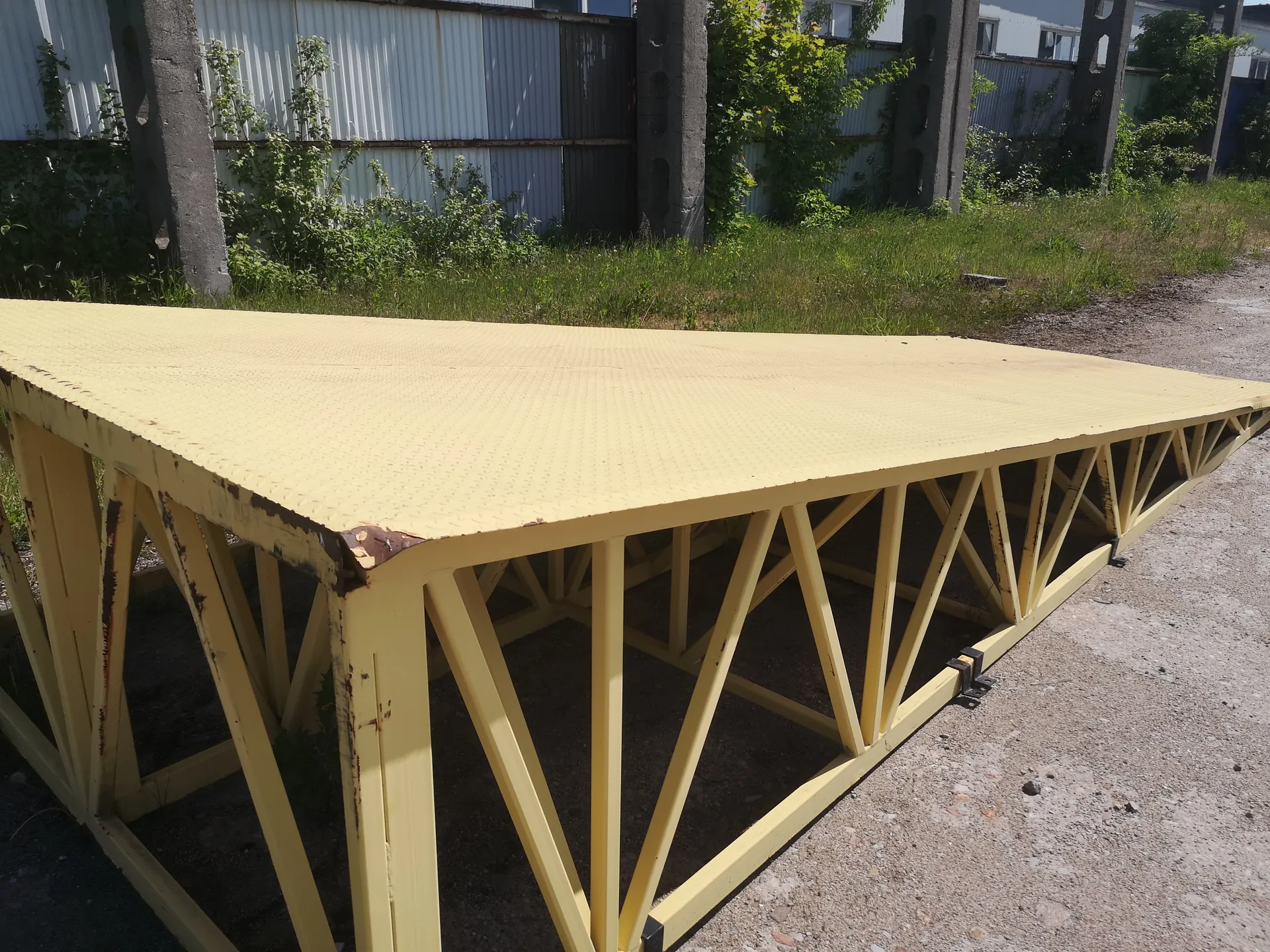 Duża żółta metalowa konstrukcja z płytką o teksturze przeciwpowrotnej i metalowymi wspornikami w kształcie trójkątów. Na krawędziach widoczna rdza. W tle trawa, metalowy płot, betonowe filary i budynki przemysłowe. Podłoże z żwiru i ziemi.