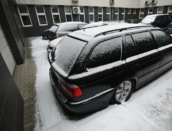 Zdjęcie przedstawia parking z kilkoma samochodami pokrytymi śniegiem. W pierwszym planie czarny samochód typu kombi z widocznym śniegiem na dachu i tylnych szybach. W tle budynki z oknami i klimatyzatorami, a także inne zaparkowane pojazdy. Na lewo od samochodów znajduje się chodnik z cegły, a cała scena jest pokryta warstwą śniegu.