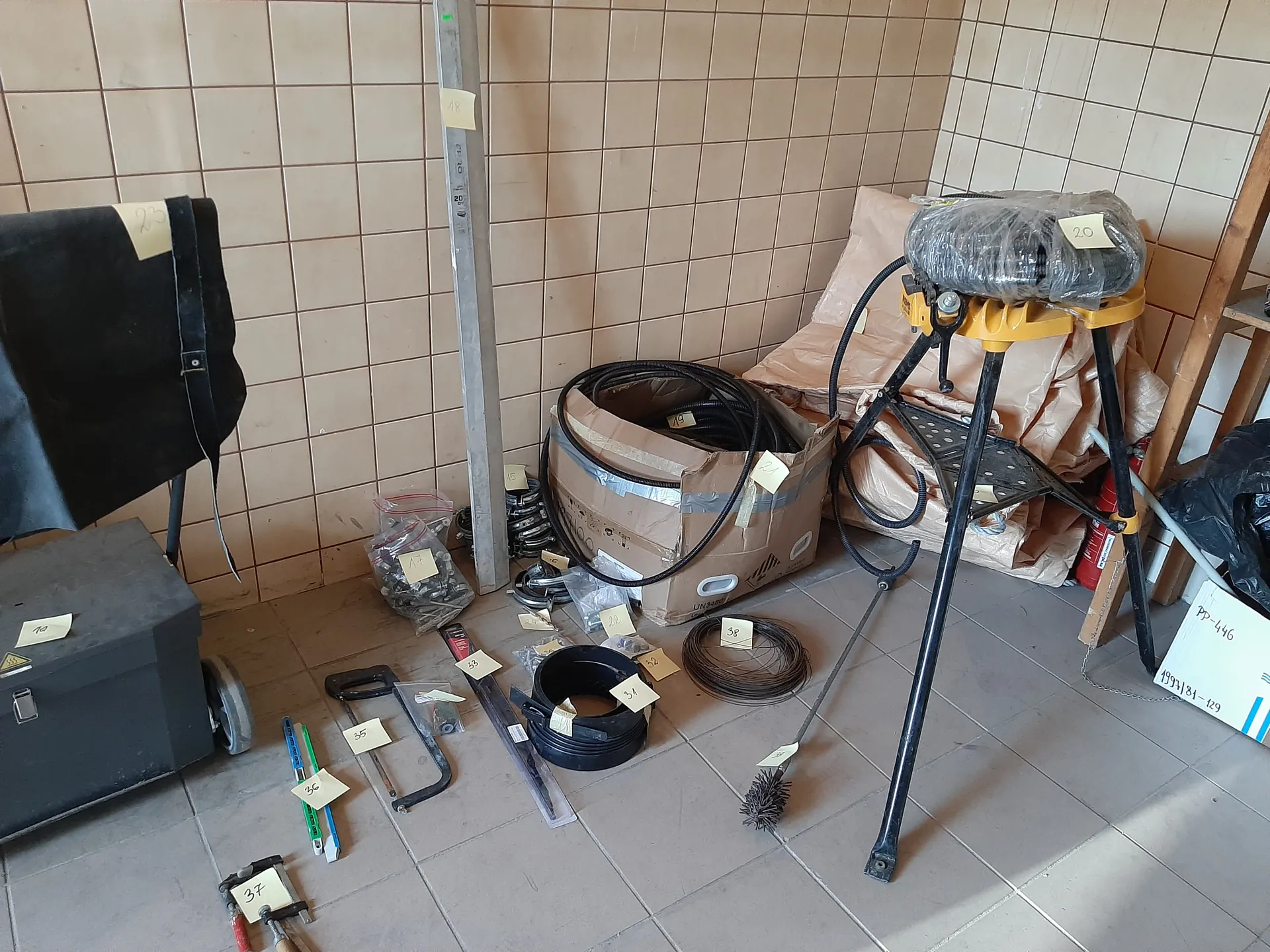 Pomieszczenie z beżowymi kafelkami na ścianach i podłodze, zawierające różne narzędzia i przedmioty. Po lewej stronie czarna skrzynia narzędziowa na kółku z etykietą 19, czarny przedmiot z etykietą 23 oraz metalowy pręt z etykietą 18. W centrum karton z czarnymi przewodami, etykietą 21 i opakowaniami z małymi częściami oznaczonymi numerami 17, 22, 31. Na podłodze: piła ręczna z etykietą 35, narzędzia z etykietami 36 i 37, zwinięty przewód z etykietą 38, szczotka i trójnóg z owiniętym przedmiotem oznaczonym 20. W tle czerwony gaśnicz, drewniana drabina oraz biały znak z napisem PP-446 i 1991/81-129. Wszystkie etykiety są czytelne, a obiekty ustawione w naturalnej orientacji.