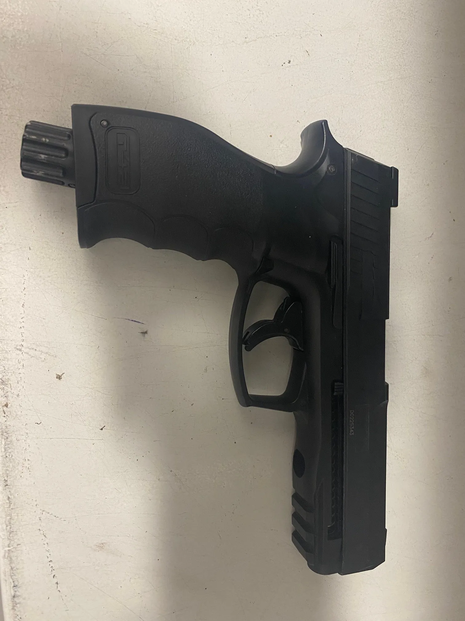 Czarny pistolet leżący poziomo na jasnej powierzchni z widocznym napisem T4E na rękojeści oraz numerem seryjnym na zamku. Pistolet ma czarną, matową powierzchnię z wyraźnymi elementami takimi jak spust, celownik i lufa z zębatą końcówką.