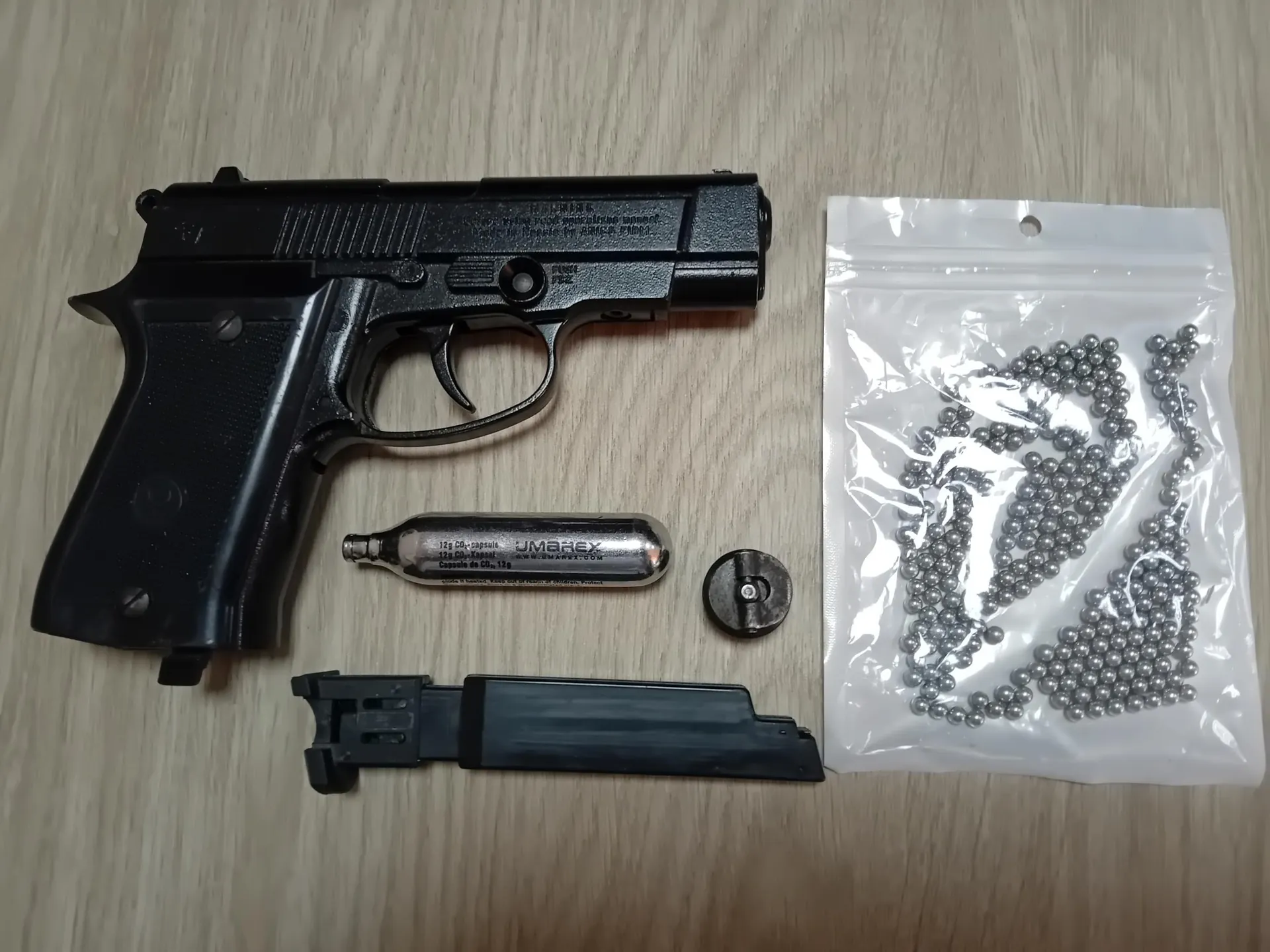 Zestaw przedmiotów ułożonych na jasnym, drewnianym blacie. W górnej części znajduje się czarny pistolet pneumatyczny. Poniżej pistoletu leży srebrny nabój z gazem z napisem 'UMBREX', mały czarny element okrągły oraz czarny magazynek. Po prawej stronie znajduje się przezroczysta foliowa torebka wypełniona metalowymi kulkami.