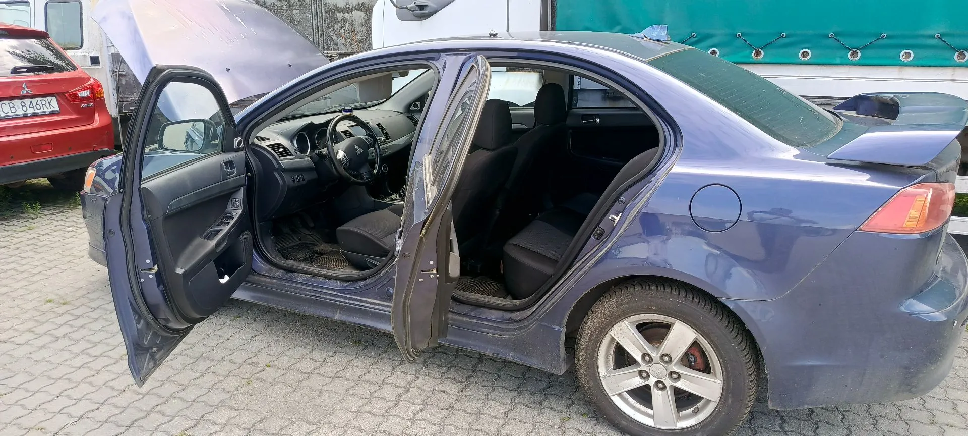 Niebieski samochód osobowy marki Mitsubishi Lancer z otwartymi drzwiami z obu stron oraz podniesioną maską silnika. Pojazd stoi na kostce brukowej. W tle widoczny jest fragment czerwonego samochodu z tablicą rejestracyjną CB 846RK oraz zielona plandeka ciężarówki.