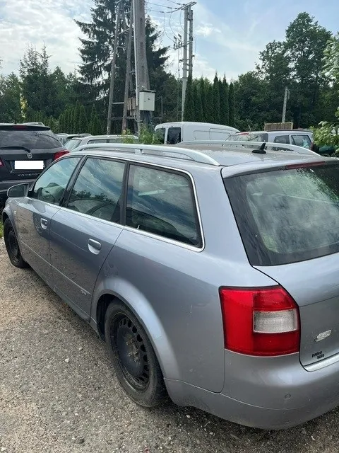 Srebrny samochód typu kombi zaparkowany na żwirowym placu. Widoczny jest tylny prawy bok pojazdu, tylna lampa oraz relingi dachowe. W tle znajdują się inne zaparkowane samochody, wysokie drzewa iglaste oraz słup elektryczny z przewodami.