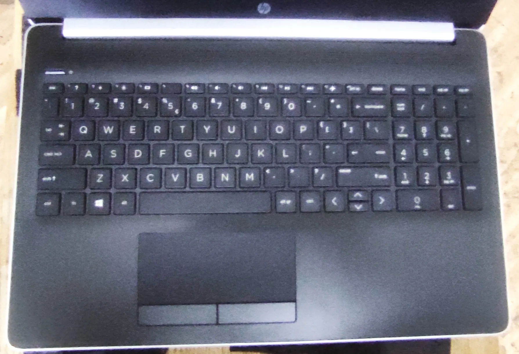 Widok z góry na czarną klawiaturę laptopa marki HP. Klawiatura posiada standardowy układ QWERTY z czarnymi klawiszami i białymi napisami. Poniżej klawiatury znajduje się duży, prostokątny touchpad z dwoma przyciskami na dole.