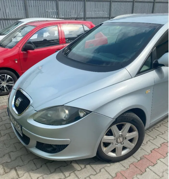 Srebrny samochód osobowy marki Seat zaparkowany na kostce brukowej. Obok niego stoi czerwony samochód. Widoczny jest przód srebrnego auta z reflektorami, grillem z logo marki oraz przednim kołem z felgą aluminiową.