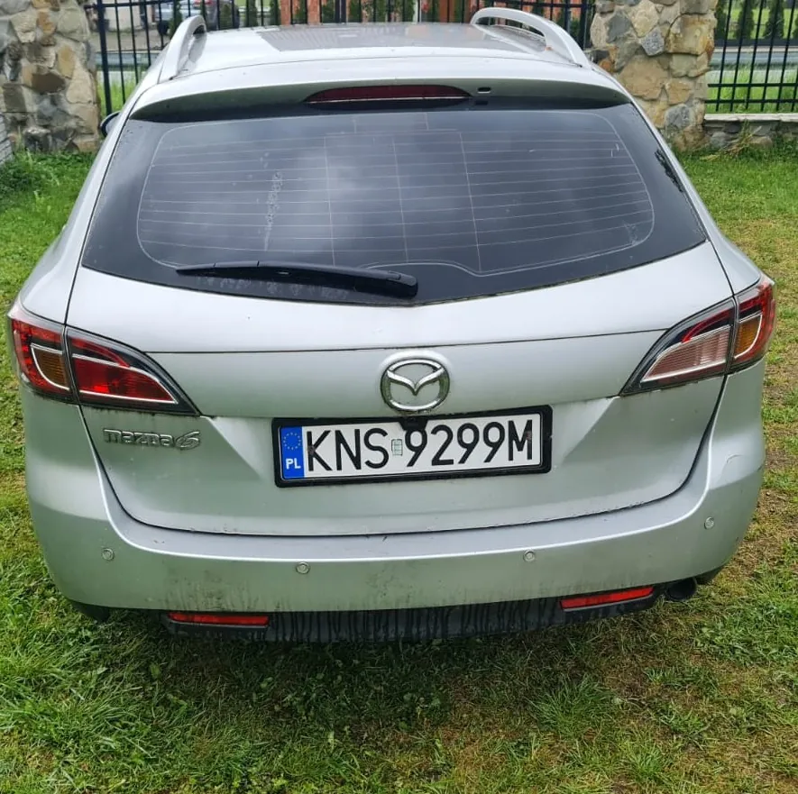 Tył srebrnego samochodu Mazda 6 z tablicą rejestracyjną KNS 9299M z kodem PL. Samochód stoi na trawie, w tle kamienna ściana i czarne metalowe ogrodzenie. Widoczne są światła tylne, logo marki, napis 'mazda 6' oraz siatka na tylnej szybie. Trawa jest zielona, a na samochodzie widoczne są ślady brudu.