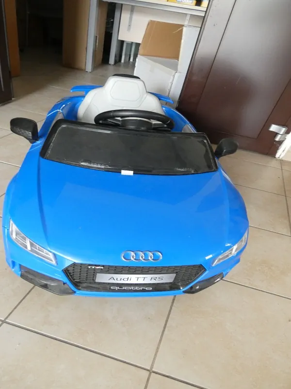 Niebieski zabawkowy samochód w stylu Audi TT RS z białym siedzeniem i czarnym kierownicą. Na przodzie widoczny emblemat Audi, tabliczka z napisem 'Audi TT RS' oraz 'quattro'. Samochód stoi na podłodze z beżowymi płytkami. W tle znajdują się kartony, brązowe drzwi i fragment pomieszczenia.