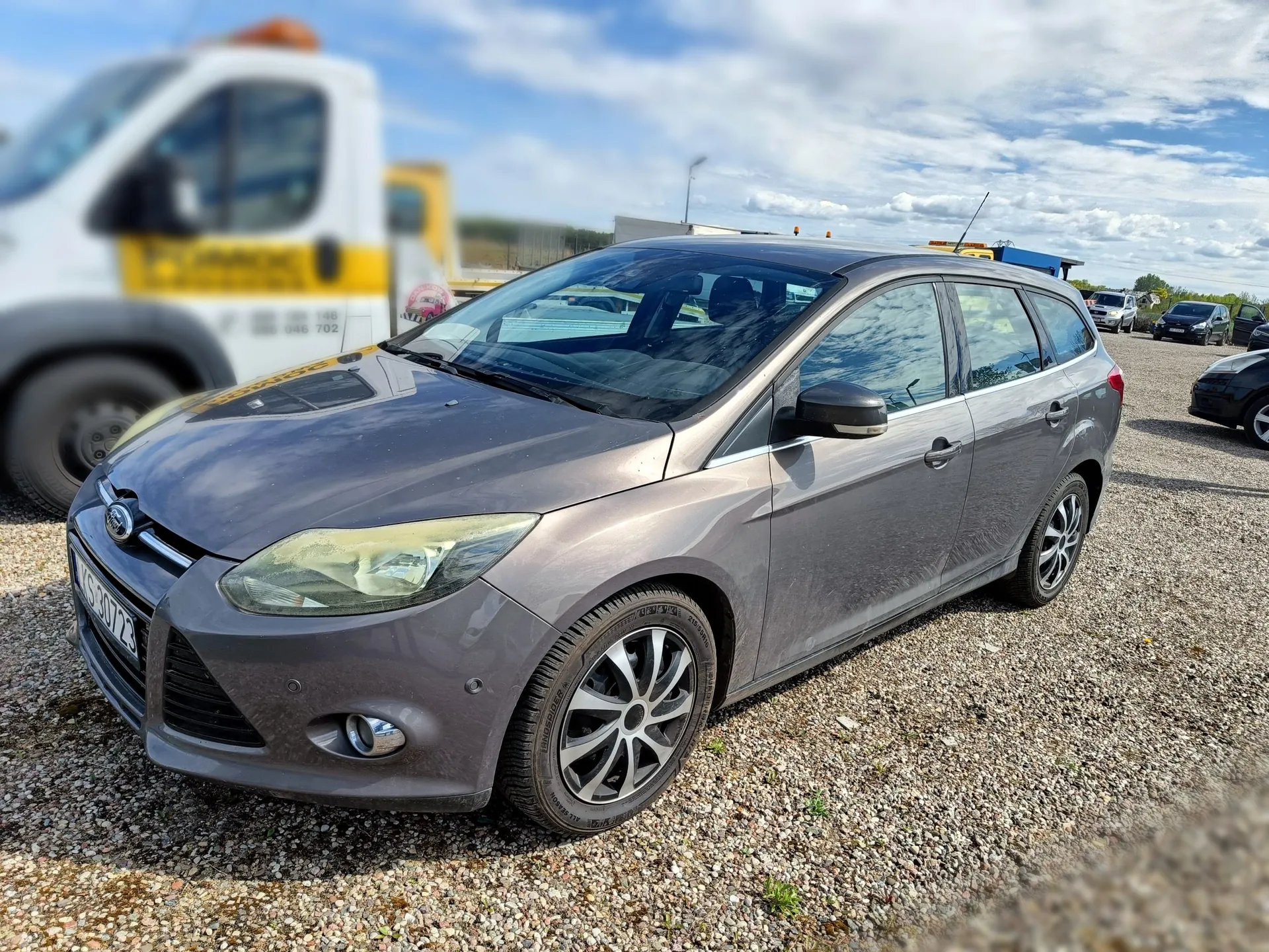 Srebrnoszary samochód osobowy marki Ford w nadwoziu kombi, zaparkowany na żwirowym placu. Auto jest pokazane z przodu i z boku. W tle widoczny jest rozmyty biały samochód dostawczy oraz inne pojazdy pod częściowo zachmurzonym niebem.