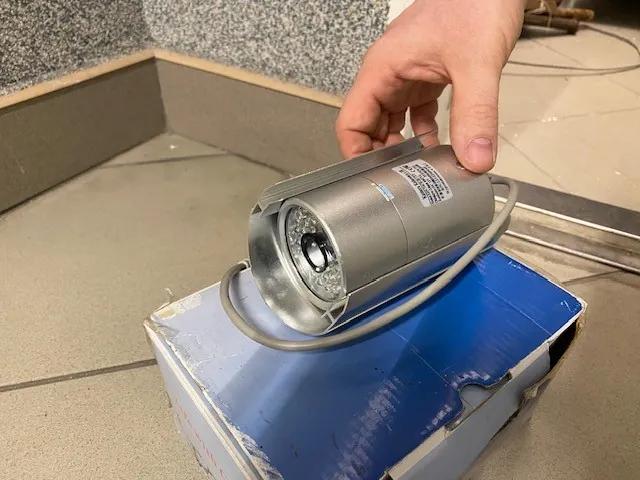 Ręka trzyma srebrny, cylindryczny obiekt przypominający kamerę monitorującą z widocznym okrągłym elementem na jednym końcu. Obiekt spoczywa na niebiesko-białym kartonie. Tło stanowi podłoga z płytek i ściana z drobnoziarnistym wzorem.