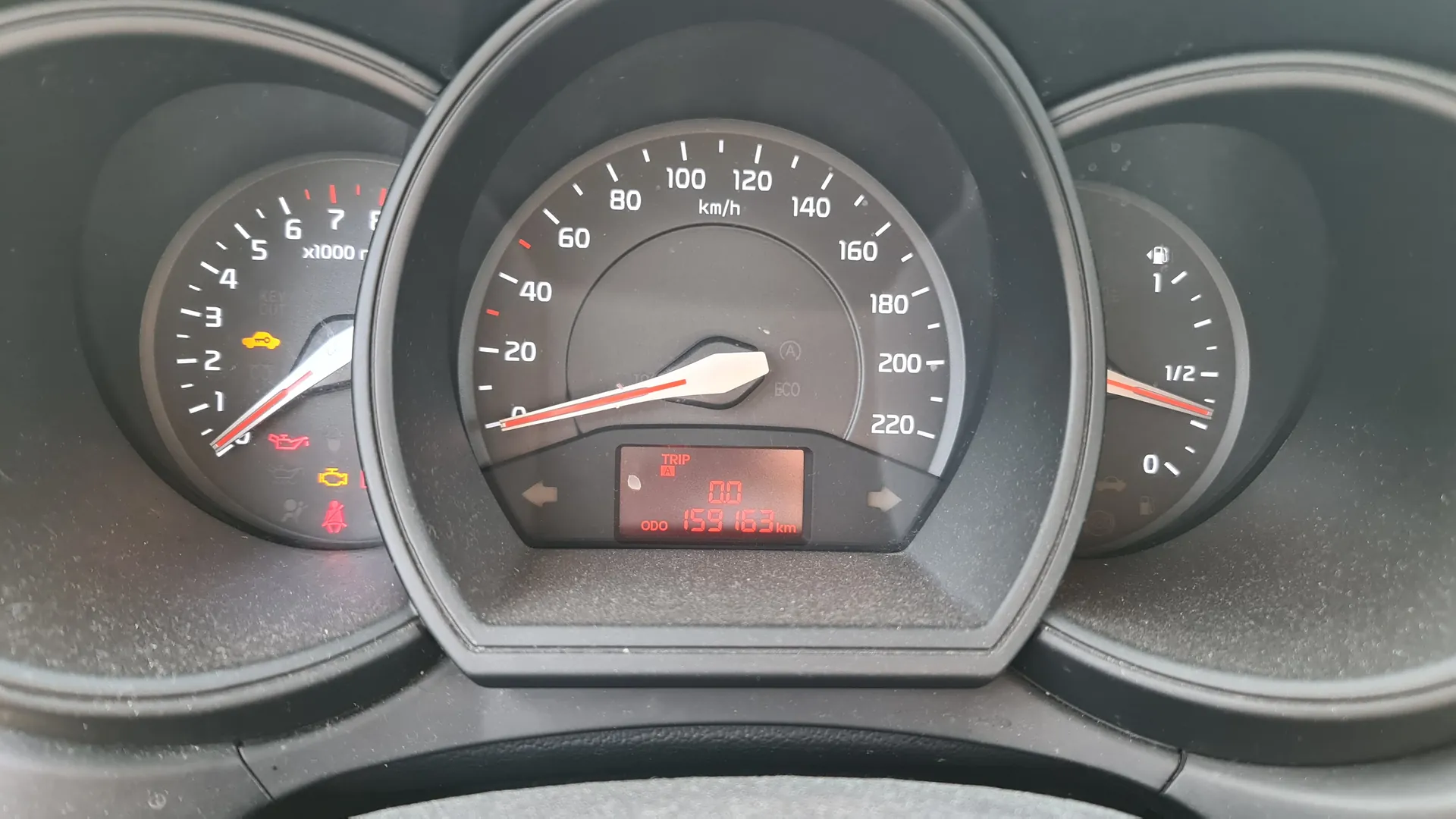 Zbliżenie na deski rozdzielcze samochodu. W centrum znajduje się prędkościomierz z zakresem do 220 km/h, wskazówka na 0 km/h. Poniżej wyświetlacz cyfrowy z napisem TRIP A, wartością 0.0 oraz ODO 159163 km. Po lewej stronie tachometr z zakresem do 7 (x1000 r), wskazówka na ok. 1, oraz ikony ostrzegawcze: żółty symbol kluczyka w samochodzie, czerwony symbol ciśnienia oleju, żółty symbol lampy silnika i czerwony symbol pasa bezpieczeństwa. Po prawej stronie wskaźnik paliwa z podziałką 0, 1/2, 1, wskazówka nieco powyżej 1/2. Na prędkościomierzu widoczny napis ECO oraz jednostka km/h.