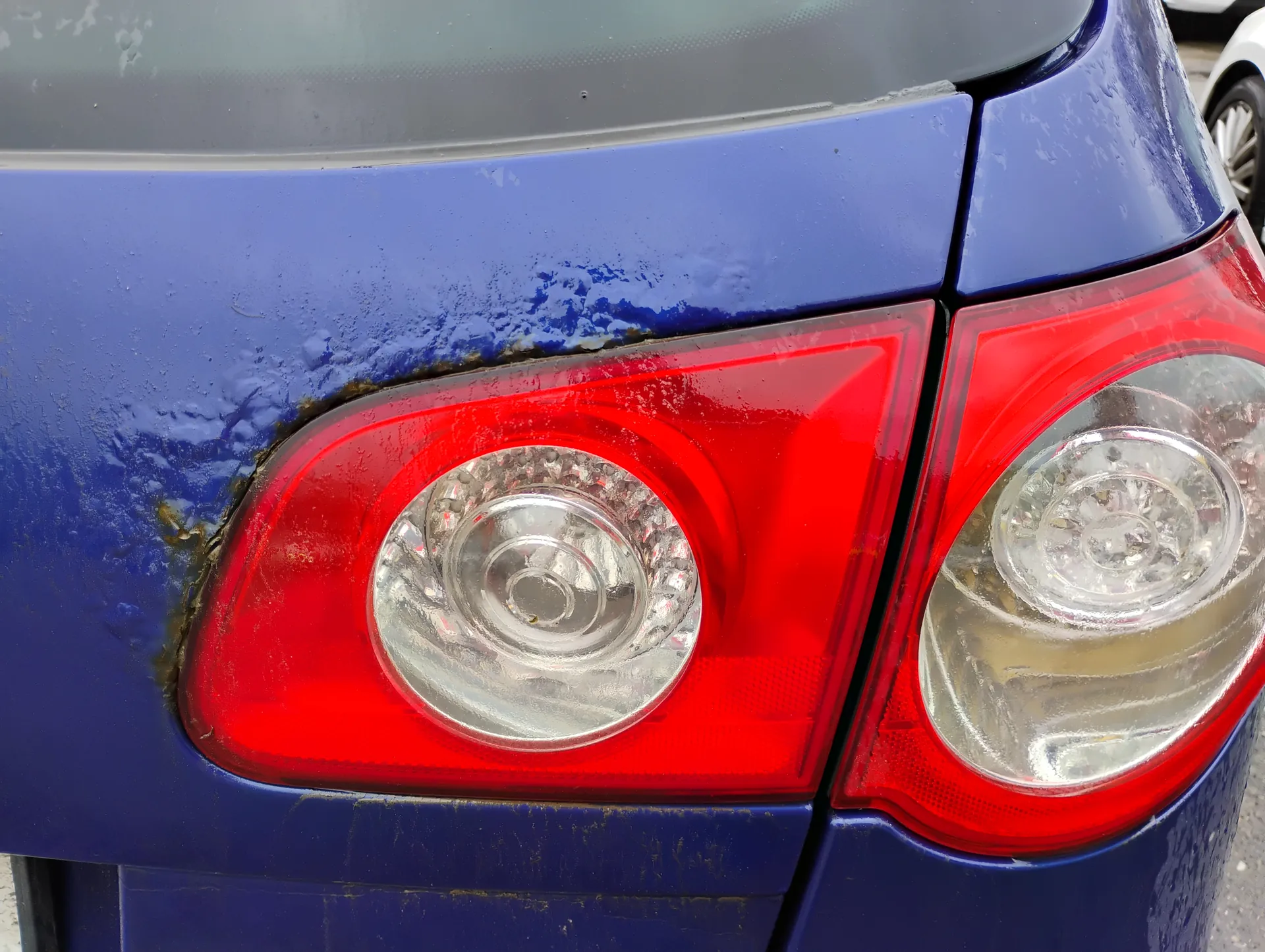 Zbliżenie na tylną część niebieskiego samochodu z dwoma czerwonymi światłami tylnymi o przezroczystych okrągłych elementach. Na lewym świetle widoczne są ślady korozji i zabrudzeń na niebieskiej karoserii. W tle częściowo widoczny biały samochód.