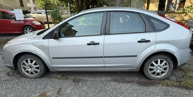 Srebrny samochód osobowy typu hatchback zaparkowany bokiem na szarym podłożu. W tle widoczny jest czerwony samochód oraz fragmenty budynków i zieleni.