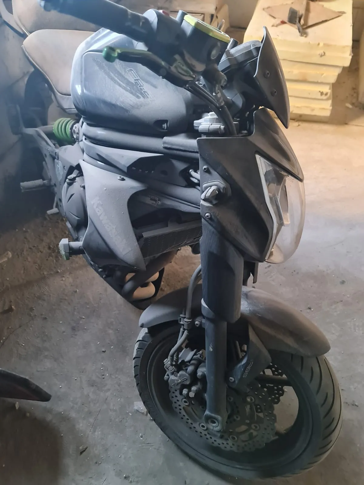 Czarny motocykl marki Kawasaki widoczny z przodu, z reflektorem, przednim kołem z hamulcem tarczowym, kierownicą z elementami sterującymi oraz widocznym napisem 'Kawasaki' na boku i 'ABS' na przednim widelcu. Motocykl znajduje się w garażu lub warsztacie z betonową podłogą i stosami drewnianych desek w tle.