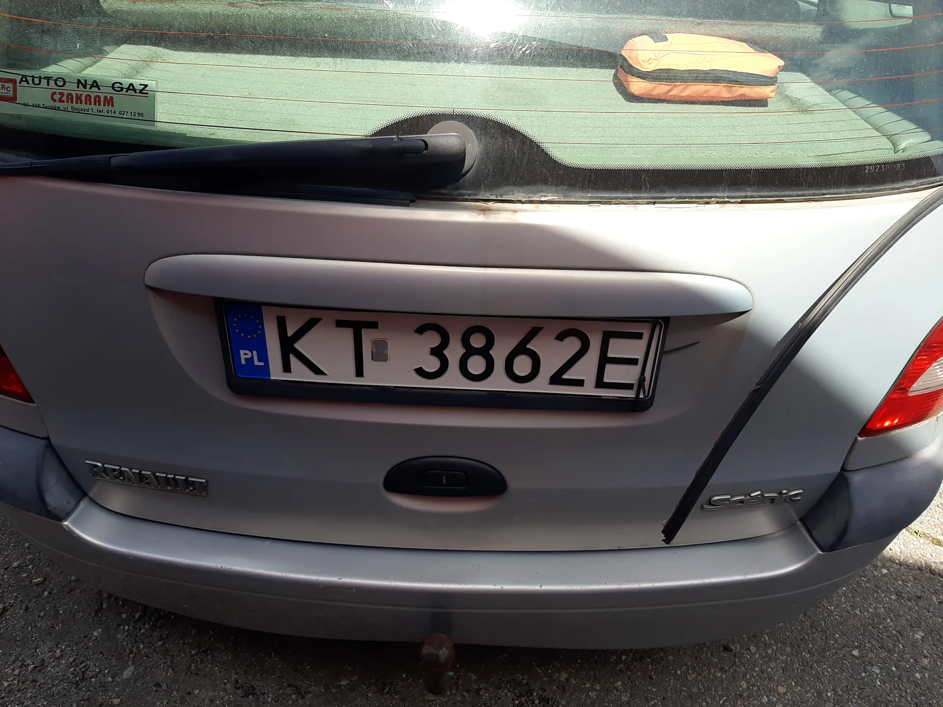 Tył srebrnego samochodu marki Renault Scénic z tablicą rejestracyjną KT 3862E (PL). Na szybie tylnej naklejka z napisem AUTO-NA-GAZ CZAKRAM oraz pomarańczowy przedmiot. Widoczne są czerwone światła tylna, uchwyt na bagażnik i fragment podłoża z drobnej kostki.