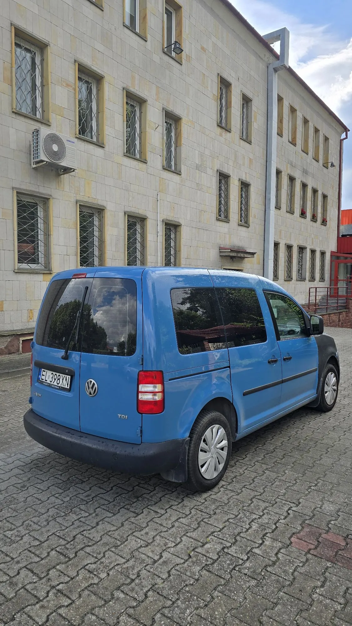 Niebieski samochód dostawczy marki Volkswagen Caddy zaparkowany tyłem na kostce brukowej przed jasnym, wielopiętrowym budynkiem z licznymi oknami zabezpieczonymi kratami. Na tylnej klapie samochodu widoczna jest tablica rejestracyjna o numerze EL 398XN oraz oznaczenie TDI.