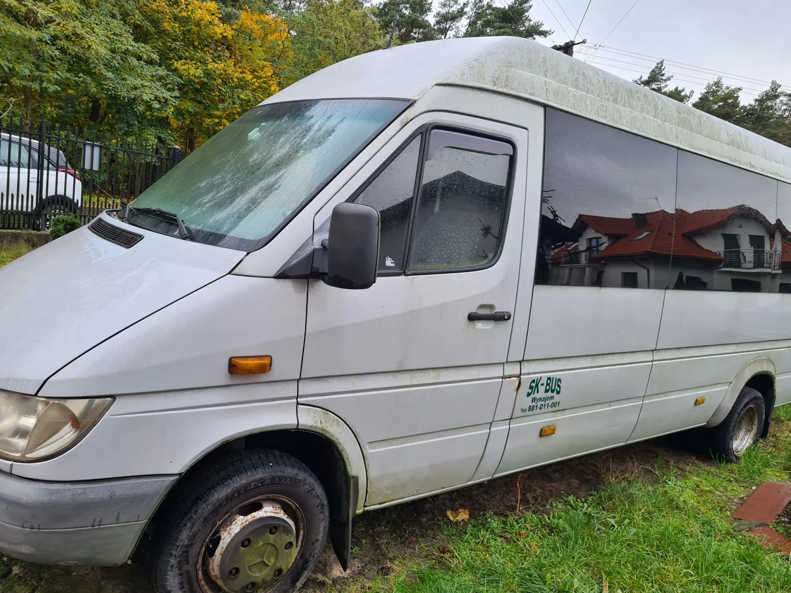 Biały furgon z napisem SK-BUS Wynajem Tel: 881-011-001 na boku, zaparkowany na trawie. Furgon ma zabrudzone koła i dolne części, przednią szybę z kroplami wody. W tle widoczne są drzewa z zielonymi i żółtymi liśćmi, czarne ogrodzenie oraz fragment białego samochodu. W oknach furgona odbijają się domy z czerwonymi dachami. Niebo jest pochmurne.