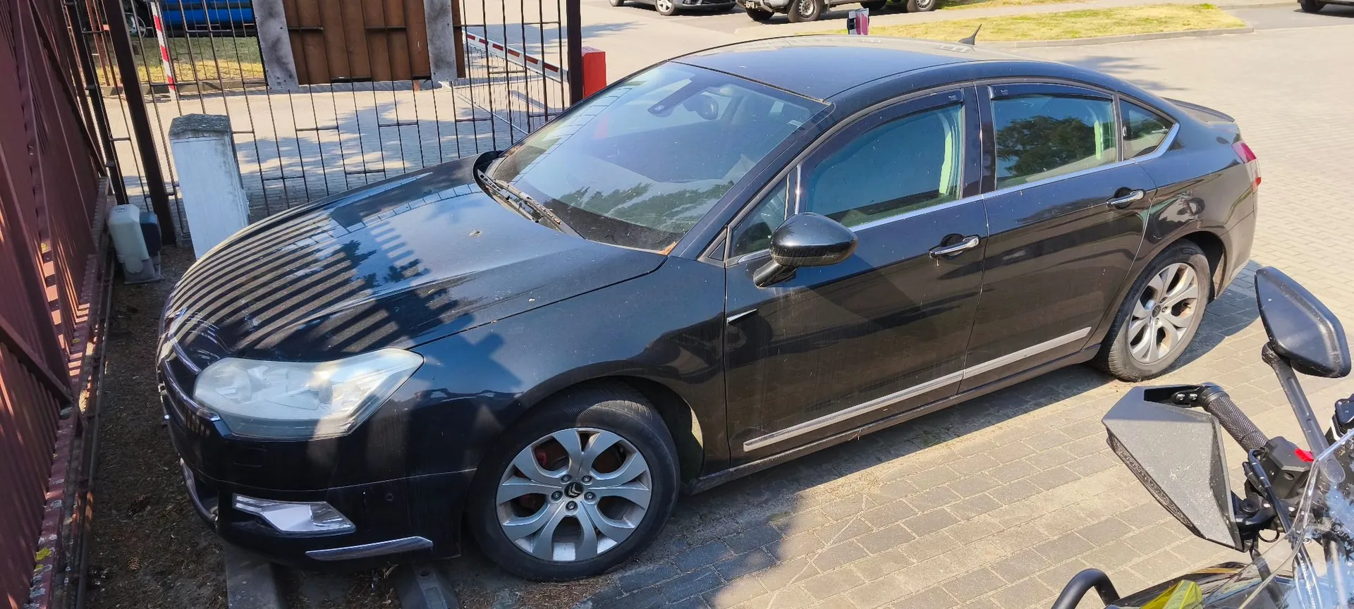 Czarny samochód osobowy typu sedan zaparkowany na kostce brukowej obok metalowego ogrodzenia. Pojazd jest widoczny z perspektywy trzech czwartych przodu, z lewej strony. W tle widać fragmenty chodnika i inne zaparkowane samochody.