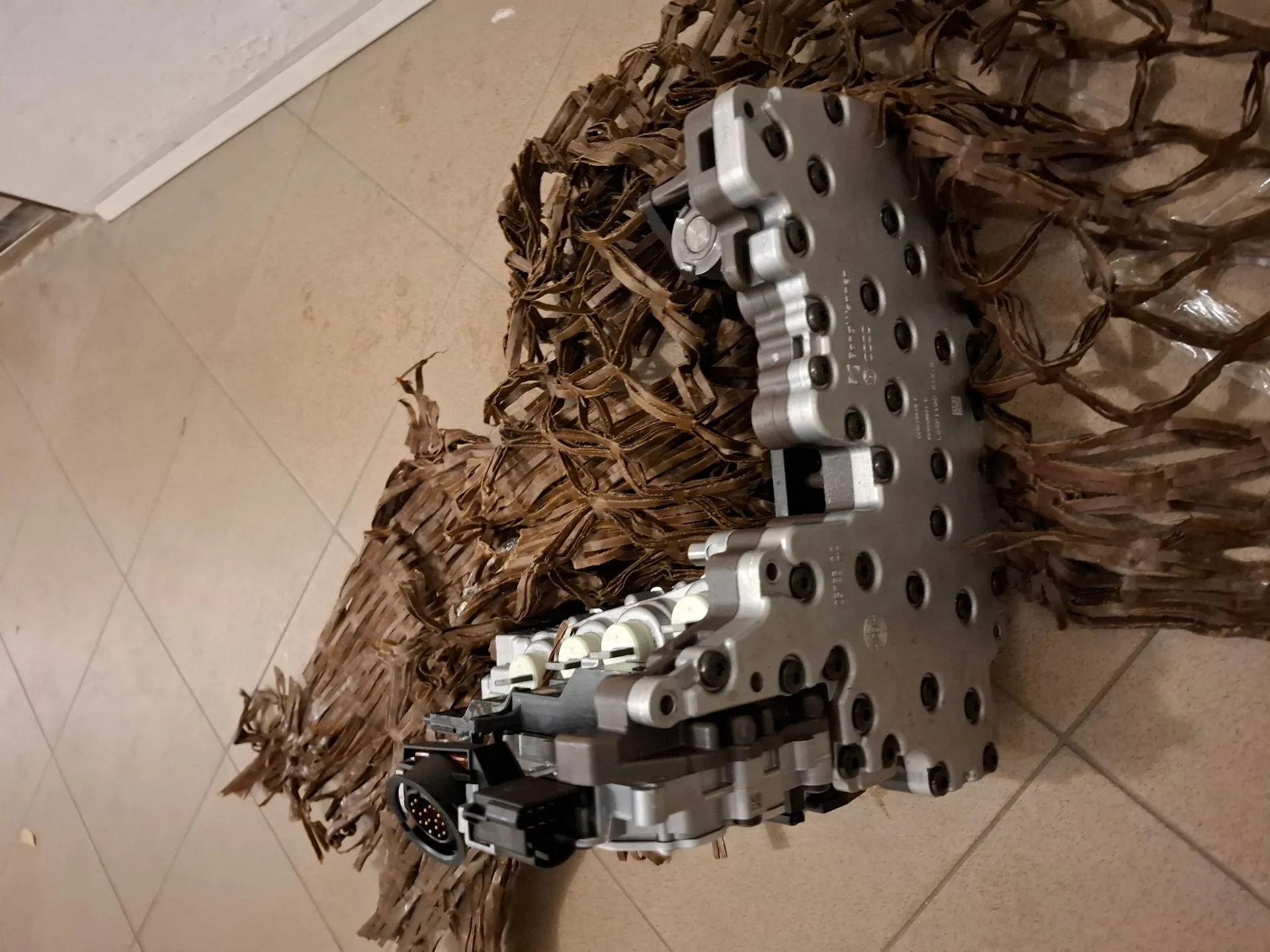 Srebrny, metalowy element mechaniczny o złożonej konstrukcji z wieloma otworami montażowymi i złączem elektrycznym, leżący na brązowym, pogniecionym papierze ochronnym na beżowej płytce ceramicznej.