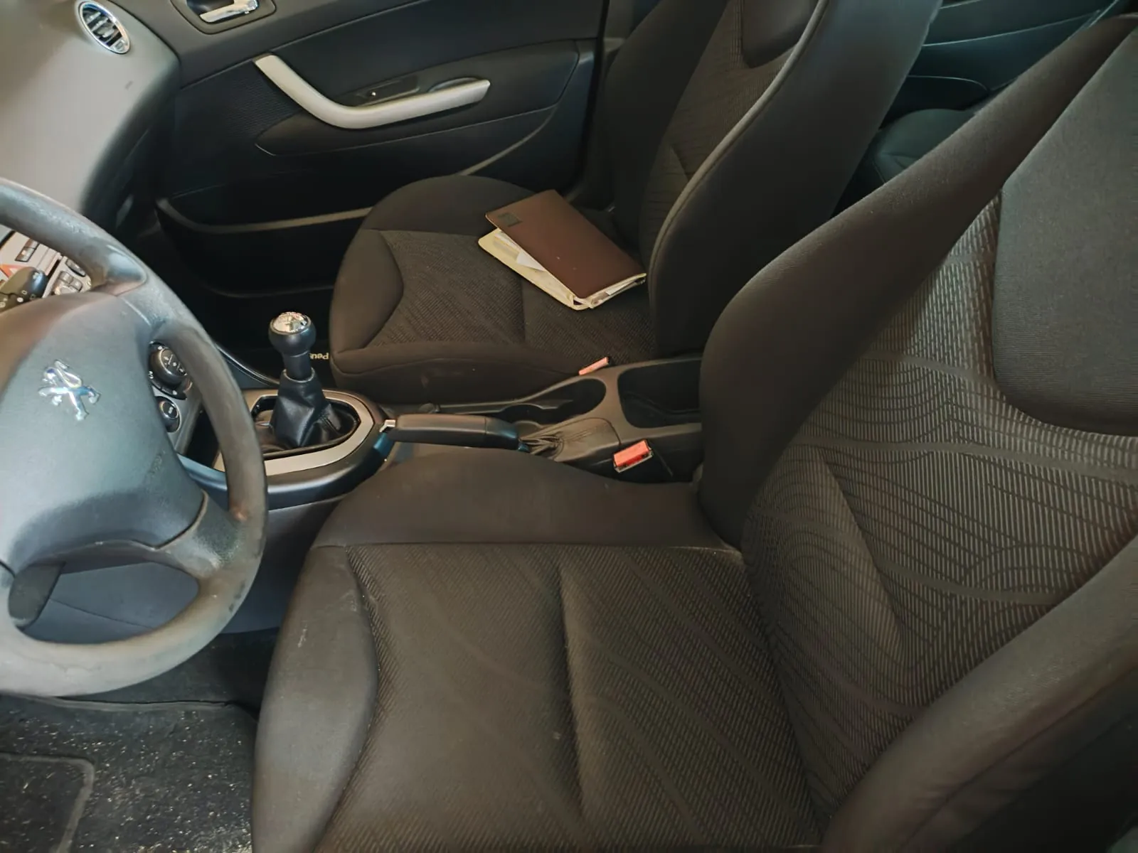 Wnętrze samochodu z widokiem na przednie fotele z ciemnej tkaniny, kierownicę z logo marki Peugeot oraz lewarek zmiany biegów. Na siedzeniu pasażera leży brązowy folder z białymi dokumentami.