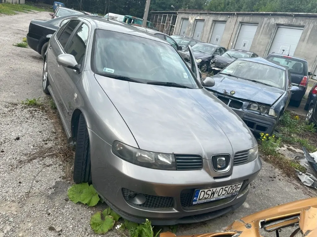Srebrny samochód marki Seat zaparkowany na nieutwardzonym placu obok innych pojazdów. Auto ma polską tablicę rejestracyjną o numerze DSW 0509E. W tle widoczny jest szary budynek z kilkoma bramami garażowymi oraz zieleń.