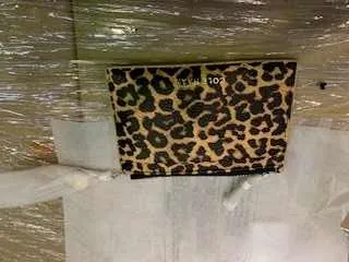 Pionowo ustawiony portfel w panterkowym wzorze z czarnymi i brązowymi plamami. Po lewej stronie portfela widoczny pionowy napis 'COACH'. Tło stanowi przezroczysta folia oraz białe przedmioty.